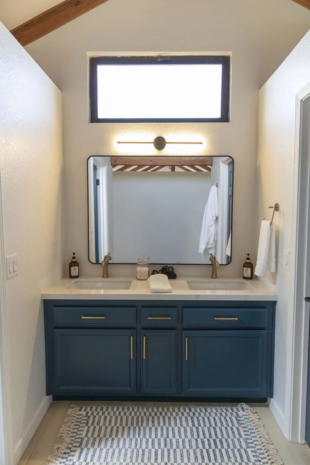 Bathroom vanity with double sinks, blue cabinet, large mirror, window above, wall-mounted light fixture, soap dispensers, towels, and decorative items.