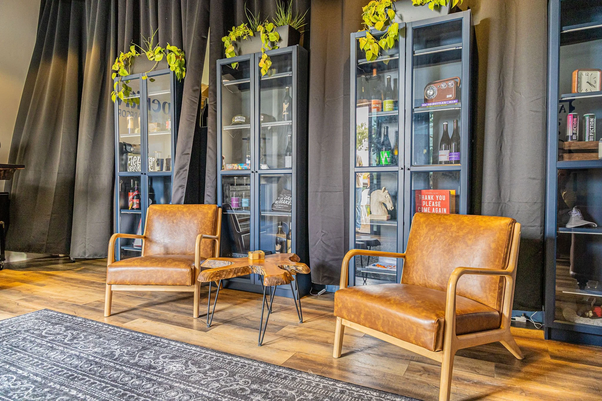 A cozy seating area with two brown leather armchairs, a wooden coffee table, and glass display cabinets filled with bottles, books, and decorative items, set against dark curtains and a wooden floor.
