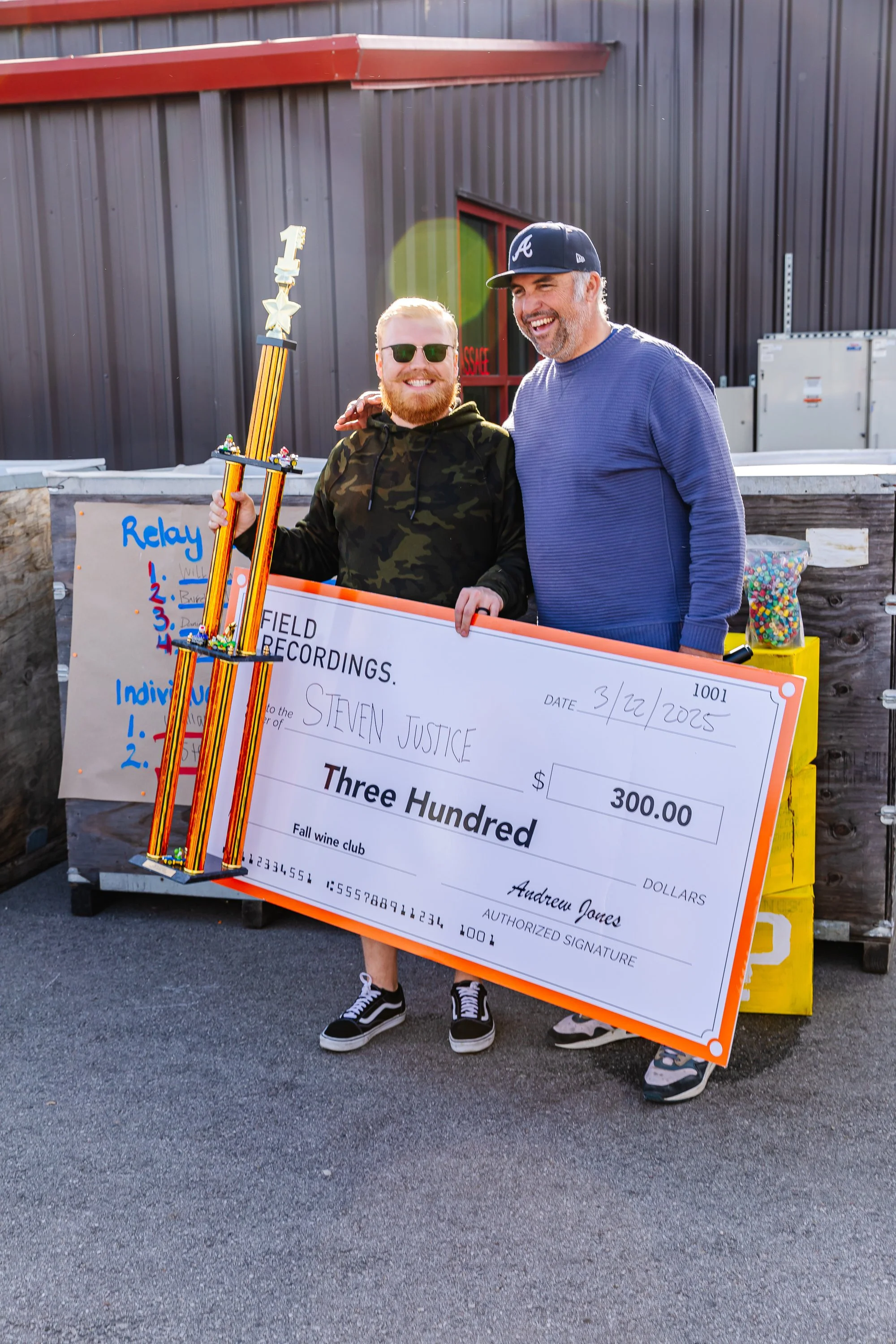 Andrew smiles and laughs with a club member, who's holding a giant trophy and a check for $300 (a complimentary club membership). They're celebrating after the Super Gnario tricycle races.