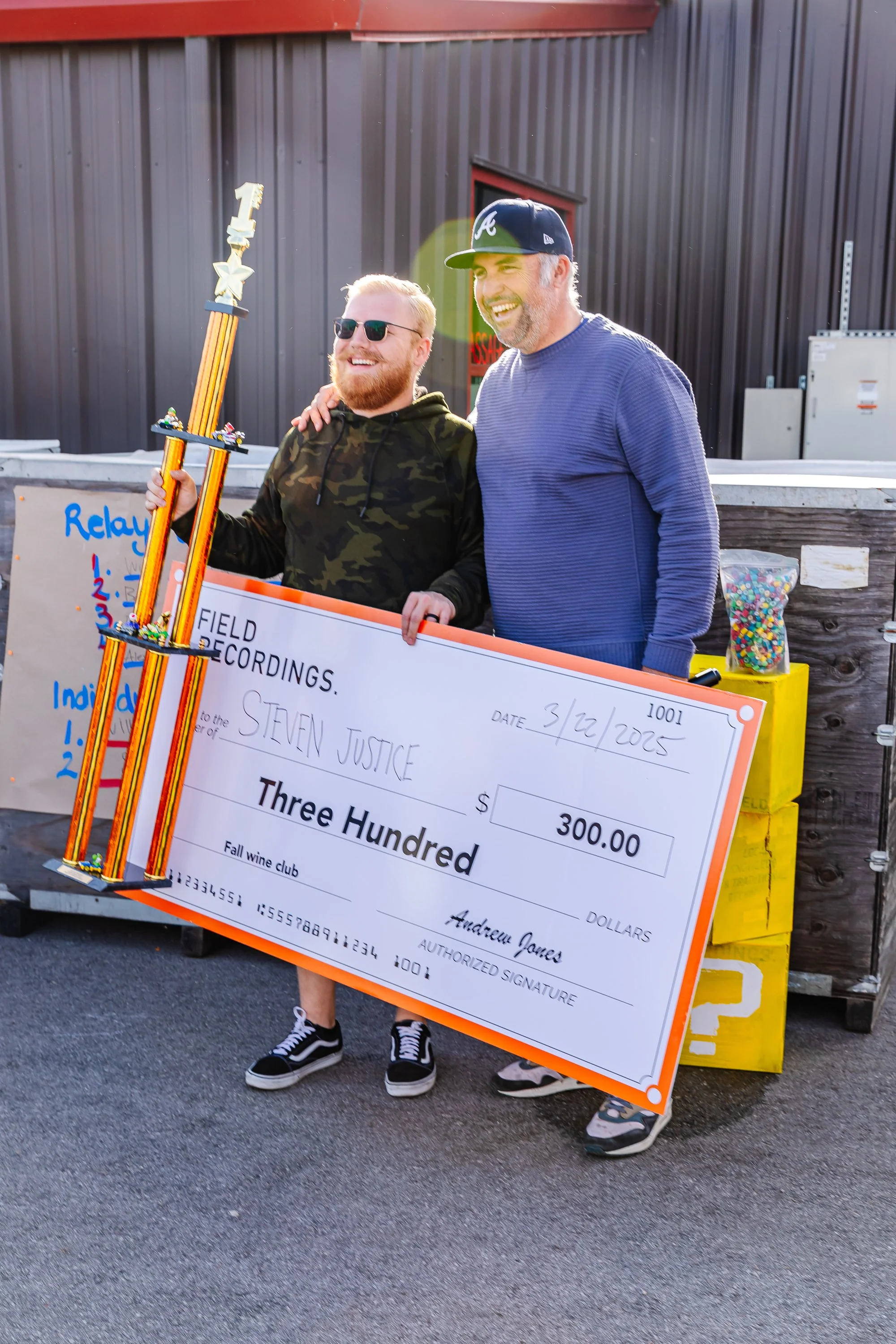 Andrew stands and laughs with a club member as they hold up a big trophy and giant check after winning a wine club competition.