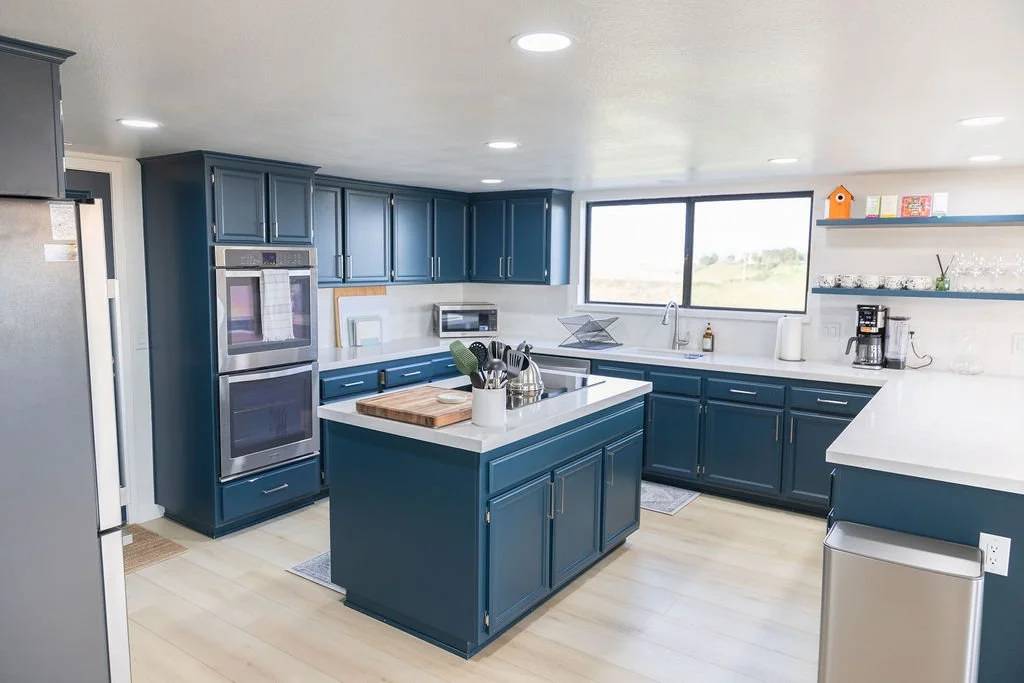 Modern kitchen with navy blue cabinets, white countertops, stainless steel appliances, and a large window providing natural light.