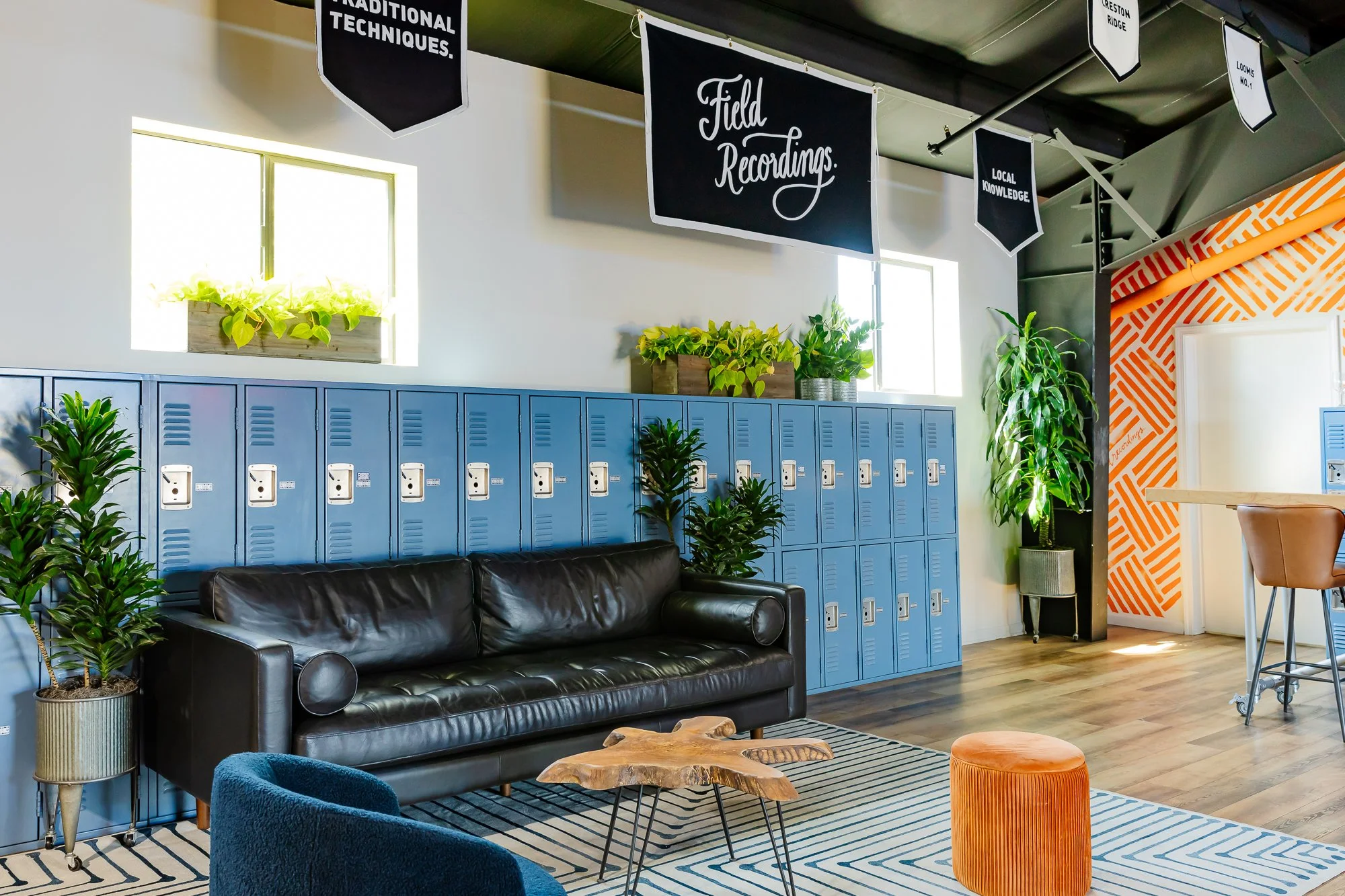 Interior of the Field Recordings tasting room with blue lockers, black leather sofa, green plants, and decorative banners hanging from the ceiling, including one that says "Field Recordings".