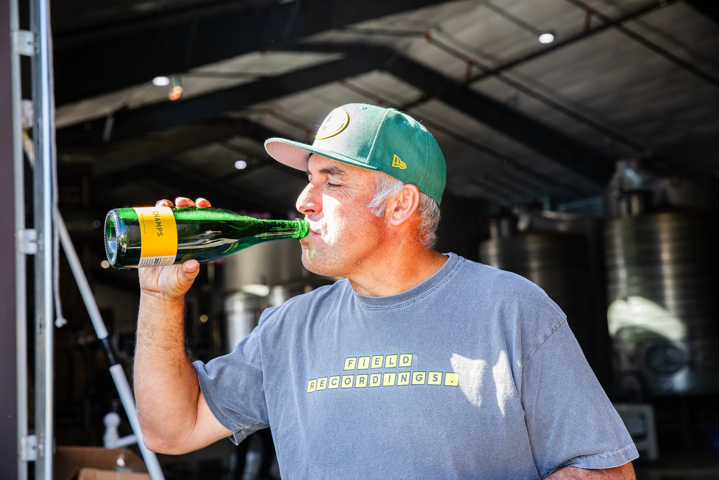 Andrew chugs from a bottle of Champs, a sparkling wine. He's wearing a gray tee and a green cap.