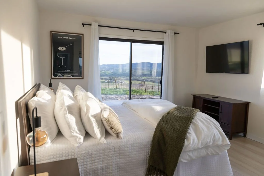 A bedroom with a large bed covered in white linens and multiple pillows, facing a window with a scenic view of fields and hills. There's a dark wood dresser and a wall-mounted TV, with a nightstand holding a lamp and decorative item.