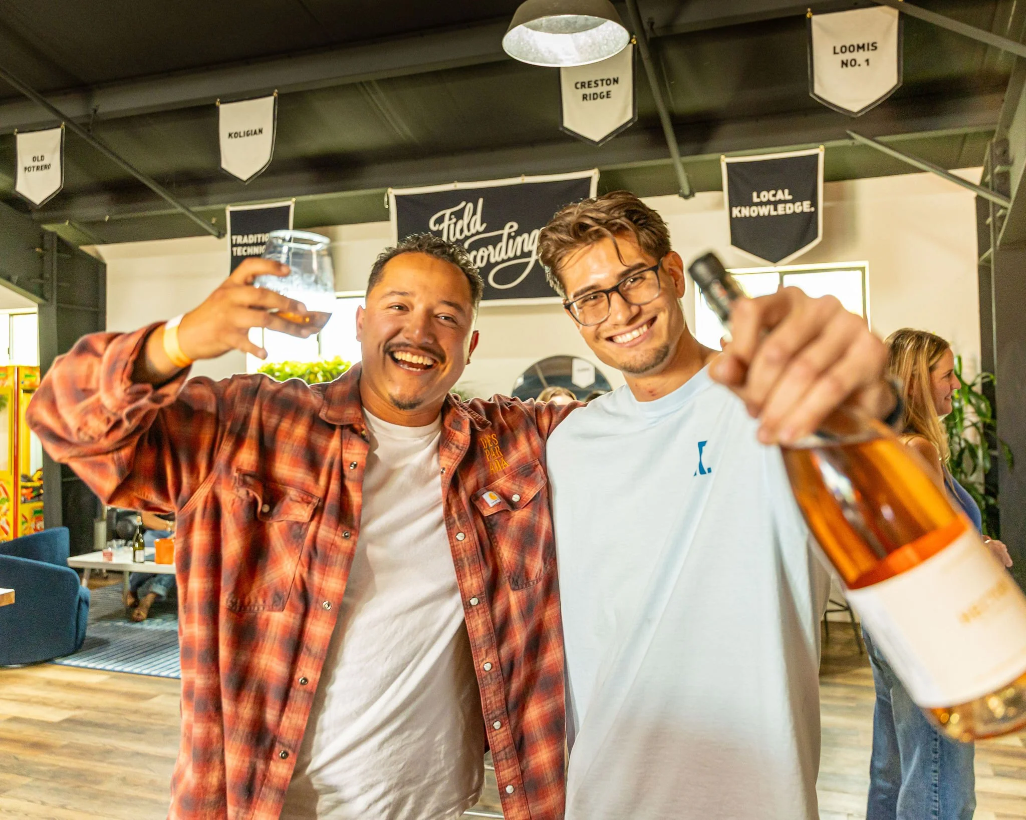 Two young men smiling and toasting with drinks in a lively social setting, with banners and people in the background.