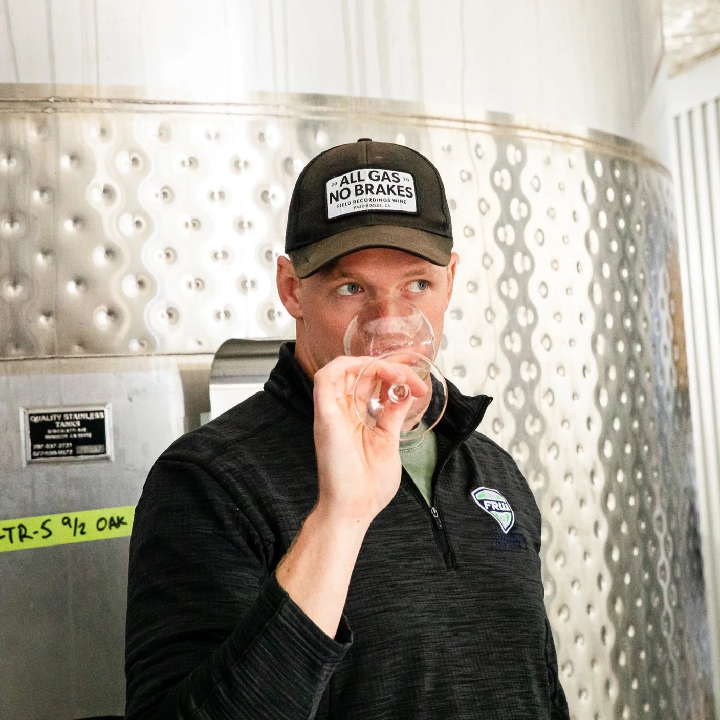 Tim stands in front of a stainless steel barrel tank and sniffs a glass of wine. He's wearing a black "All Gas, No Brakes" hat.