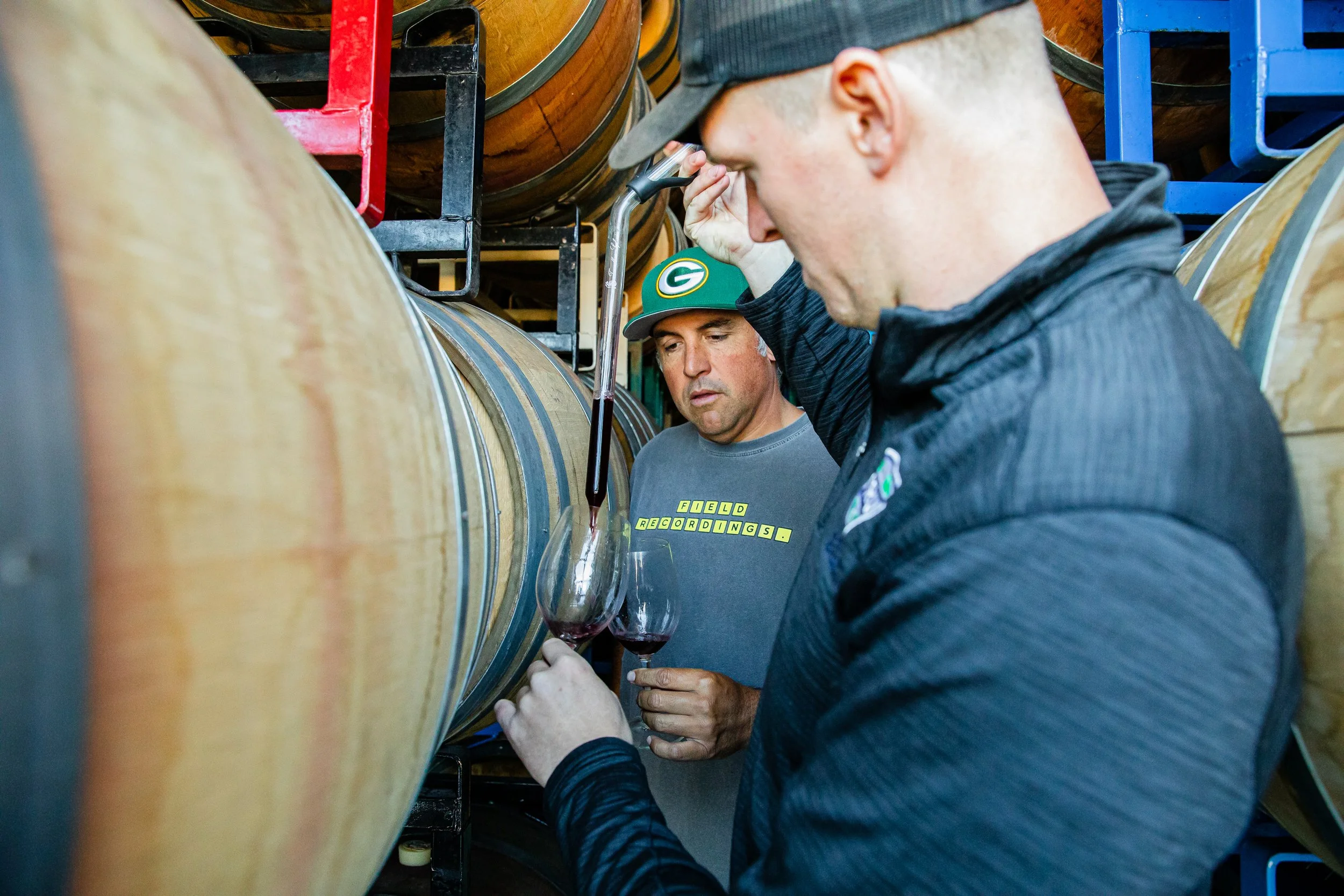 A man wearing a baseball cap and glasses inspects a glass of red wine while standing next to large wooden wine barrels in a winery.
