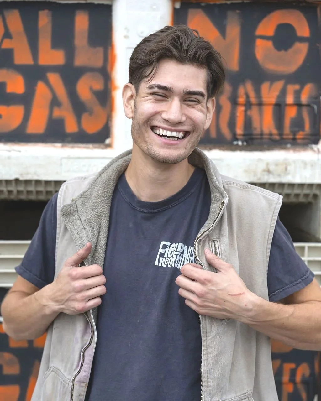 Hector stands and laughs in front of a stack of bins that read "All Gas, No Brakes."He's wearing a blue FR shirt and a khaki vest.