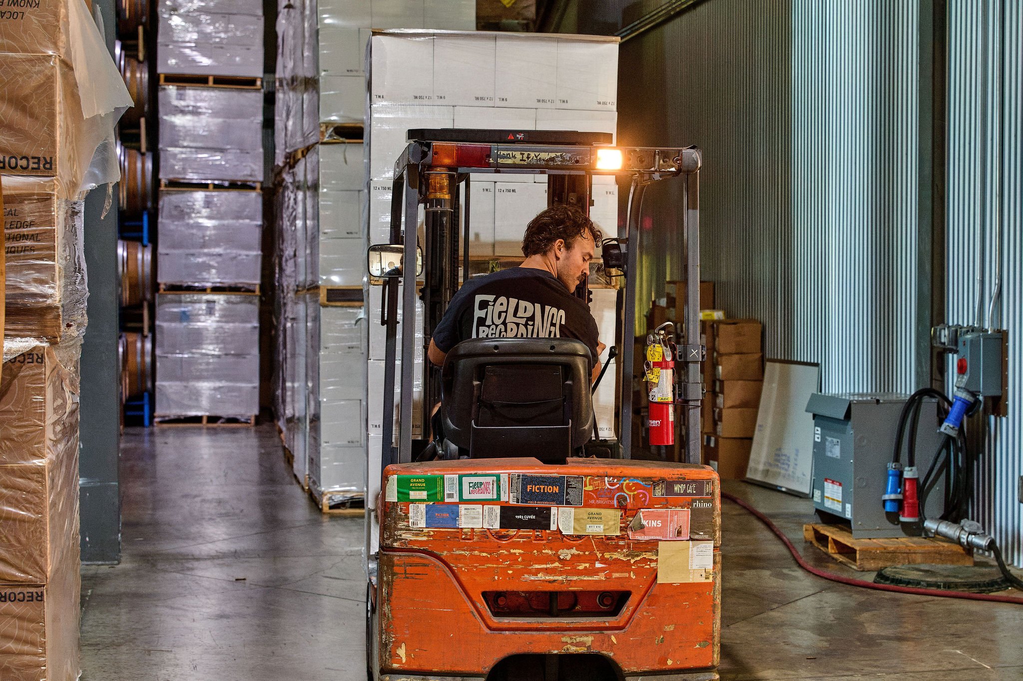 Austin turns his head back as he reverses a forklift in front of pallets of wine cases.