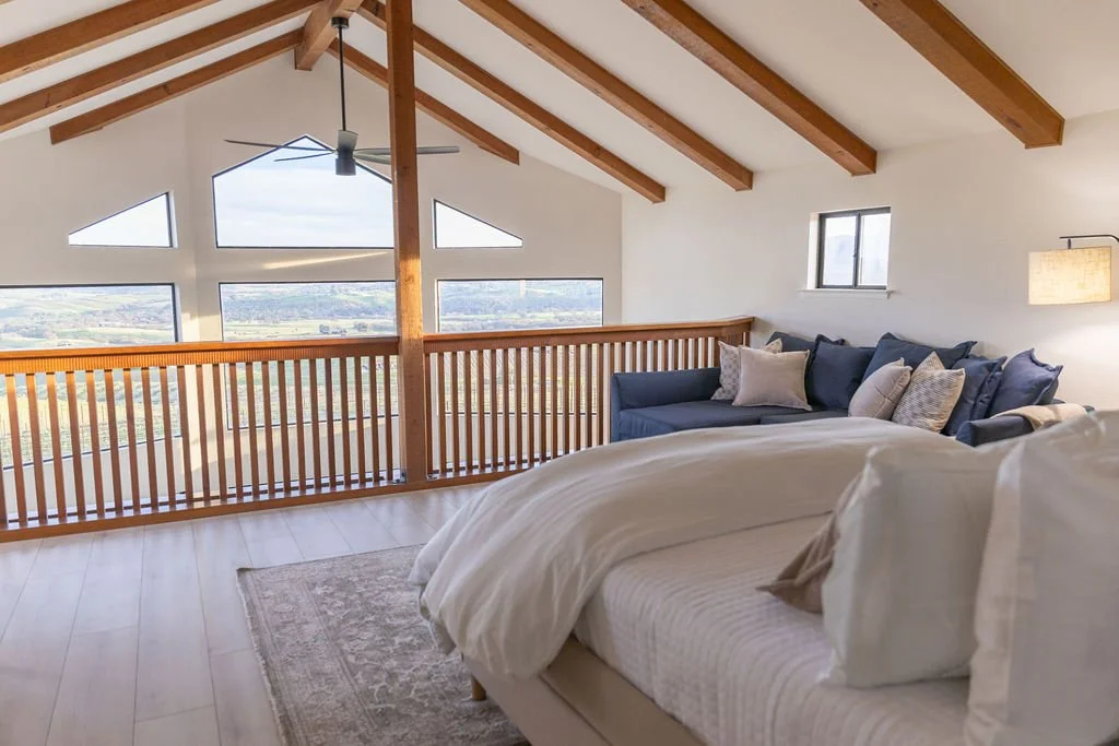 Interior of a bedroom with a wooden ceiling and large windows with mountain views, featuring a bed with white bedding and a blue couch with multiple pillows.