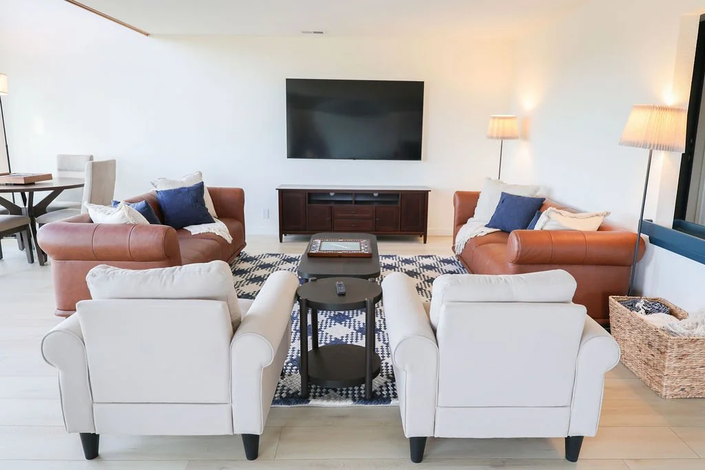 Living room with two white armchairs, two brown leather sofas, a flat-screen TV on the wall, a wooden TV stand, a black coffee table, a patterned rug, two floor lamps with beige shades, and throw pillows in white and navy blue.