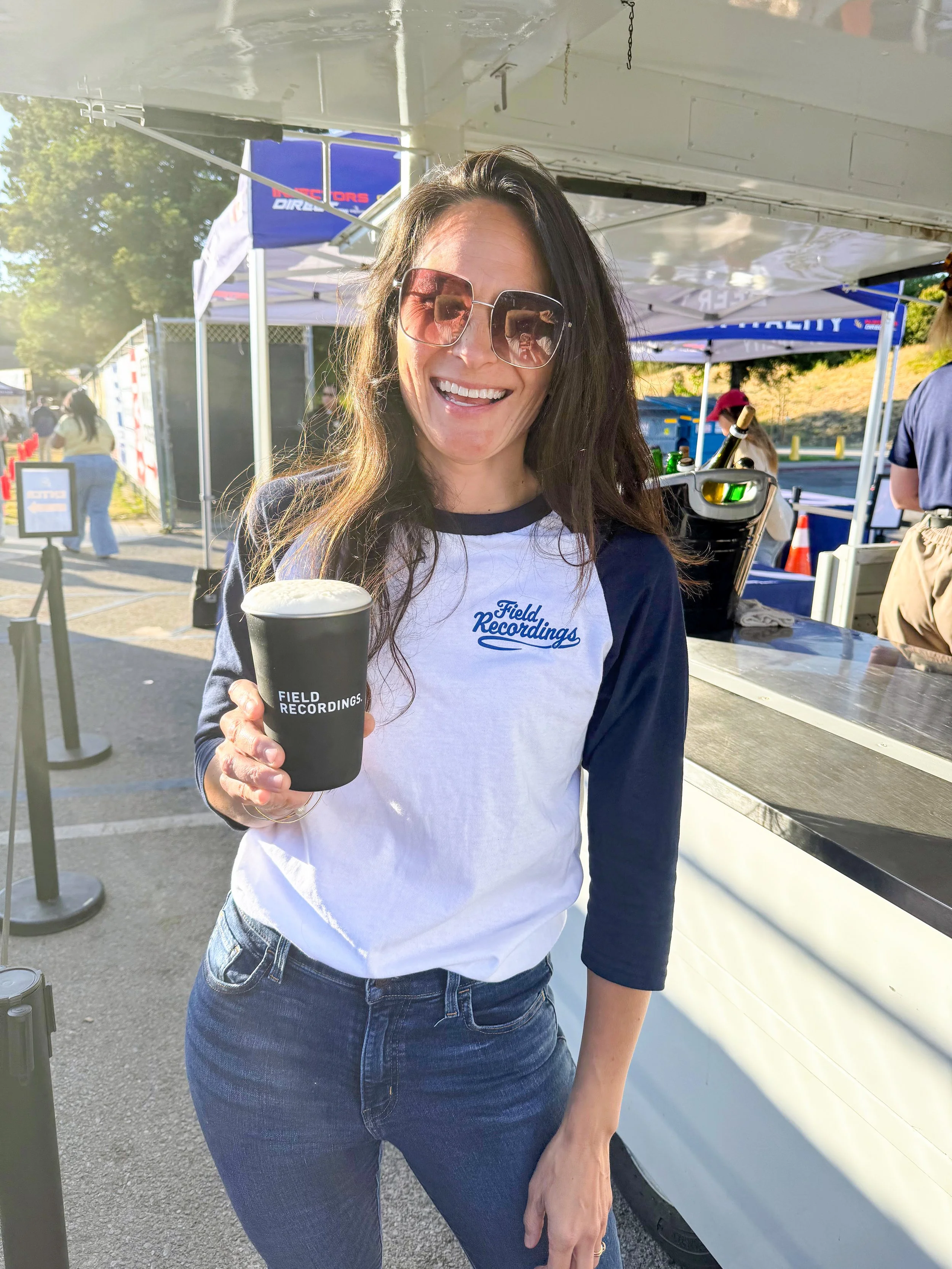 Andrea smiles and holds a Field Recordings cup full of beer. She's wearing a Field Recordings baseball tee and standing in front of the SLO Blues drink station.
