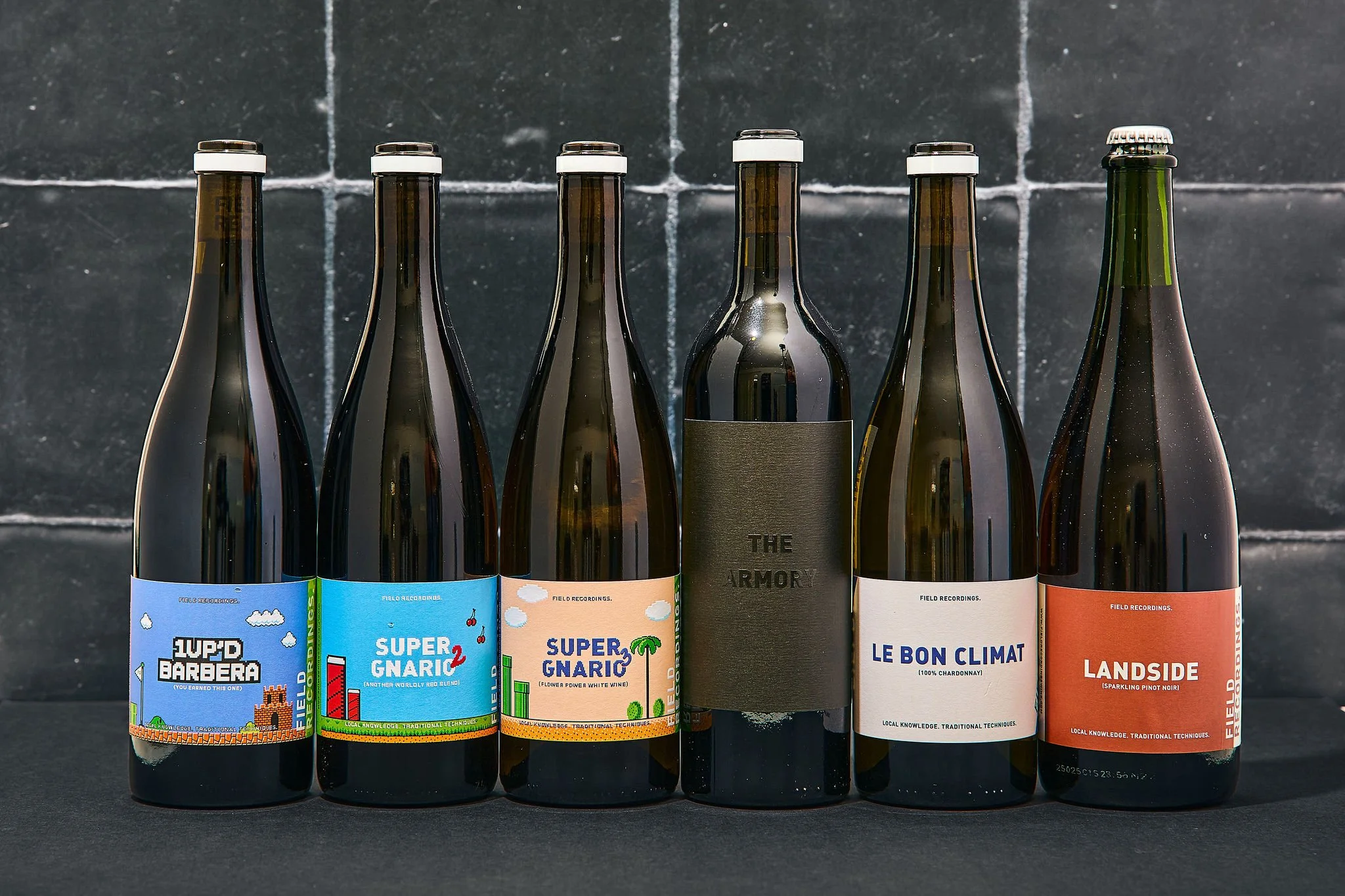 Six wine bottles lined up agianst a black tile background.