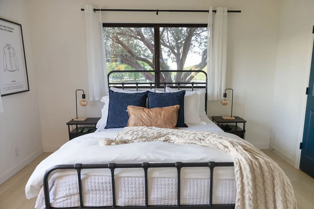 A bedroom with a black metal bed frame, white bedding, a cream knitted blanket, and four pillows (two white, two navy blue, one in front of the blue pillows in a tan color). There are two black nightstands with small lamps beside the bed. A large window with white curtains shows a tree outside.