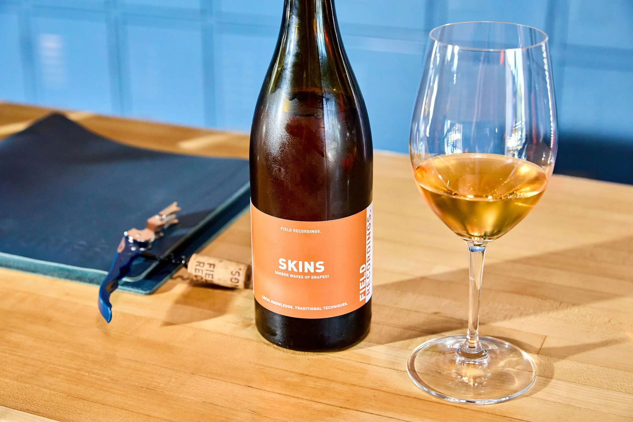 A wine bottle labeled 'Skins' and a wine glass filled with orange wine on a wooden table at the Field Recordings tasting room, with a blue background.