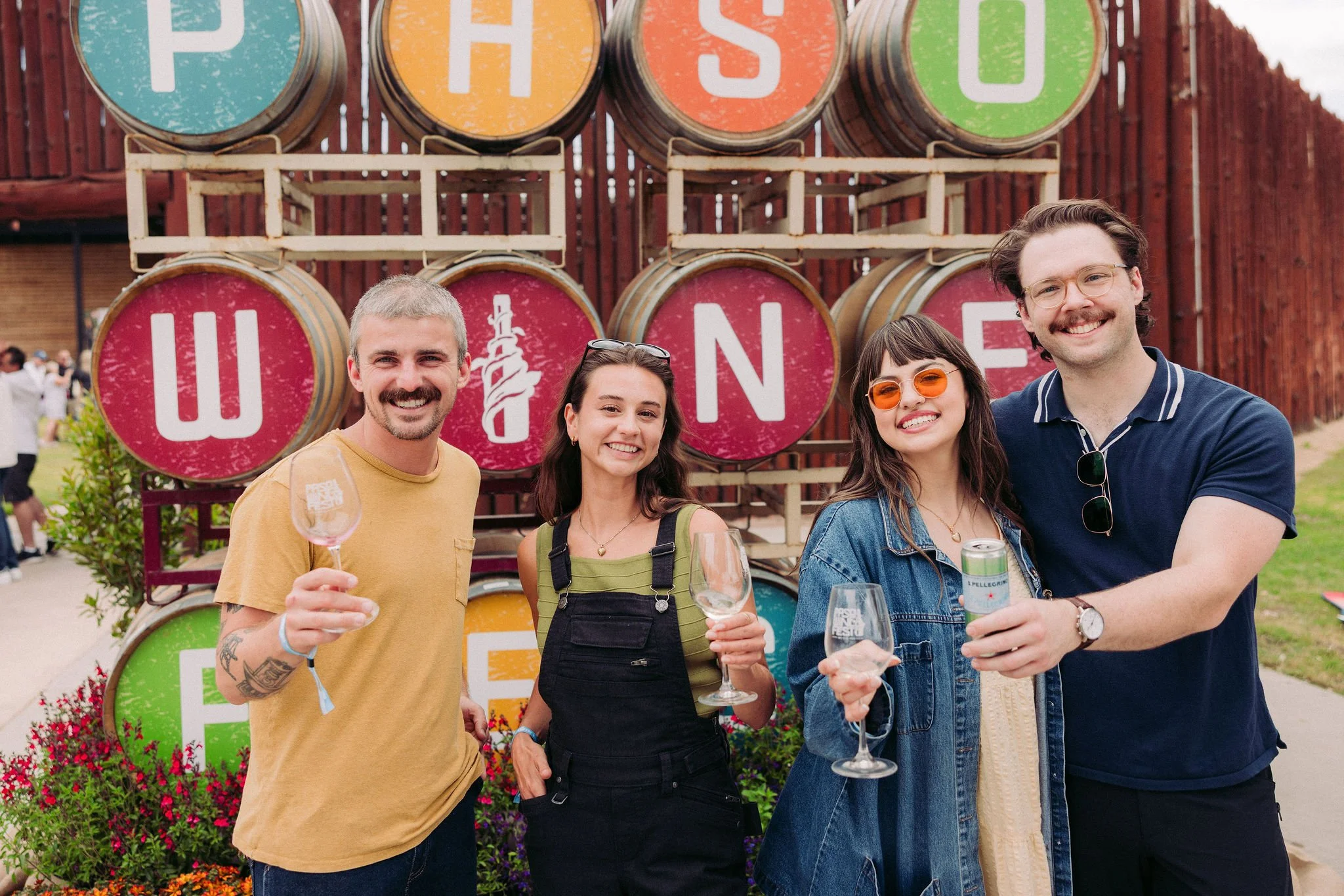 Four people smile and hold their wine glasses up in front of brightly colored barrels that read "Paso Wine"