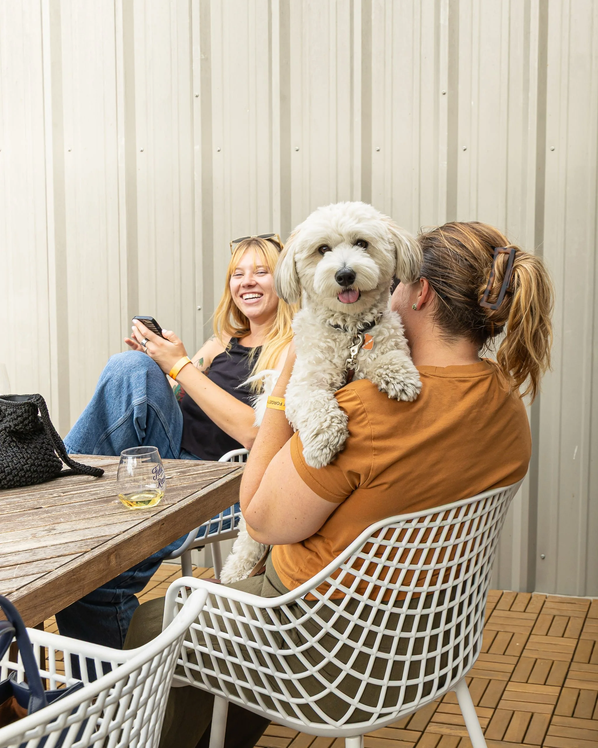 Two women are sitting at an outdoor wooden table against a metal wall, with one woman holding a small white fluffy dog on her lap. The woman on the left is smiling and holding a smartphone, the woman on the right has her back to the camera, and there is a glass of white wine on the table.