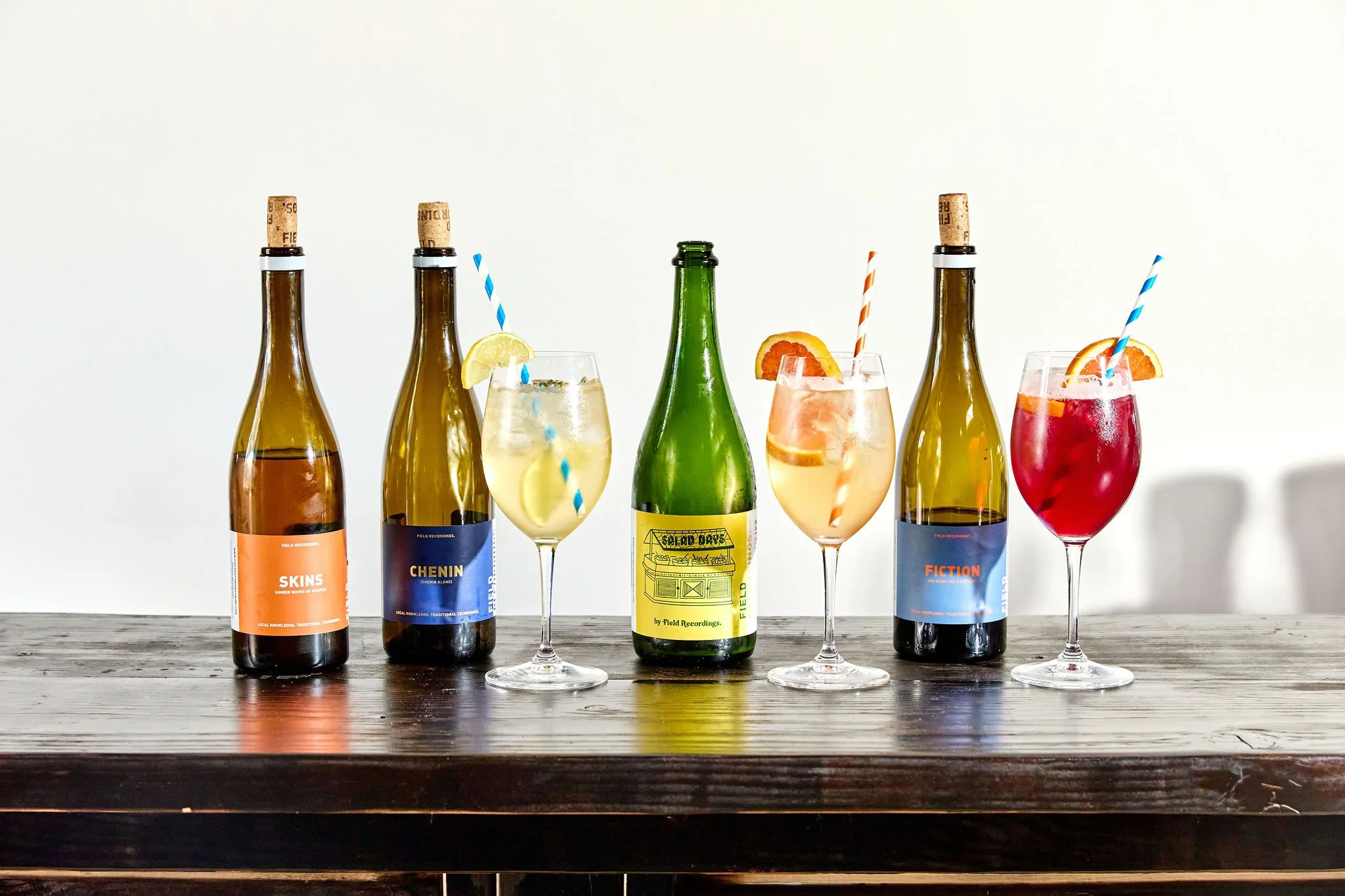 Six bottles of Field Recordings sparkling wine with colorful labels and four glasses filled with spritz cocktails garnished with lemon or orange slices and colorful paper straws, arranged on a wooden table against a white background.