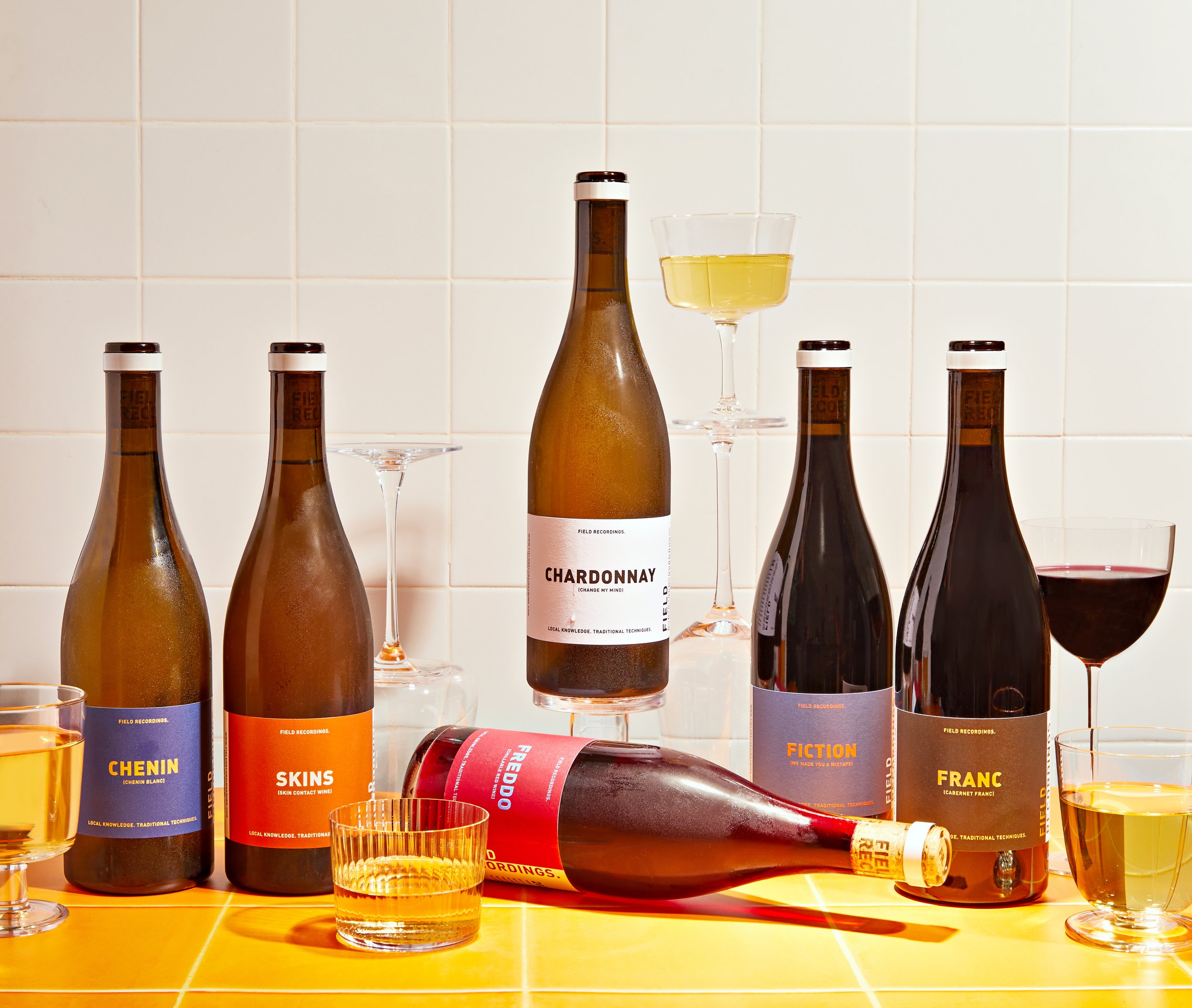 Six bottles of wine arranged on a yellow tile countertop with a white tile background. Wine glasses of various heights are stacked around the bottles.
