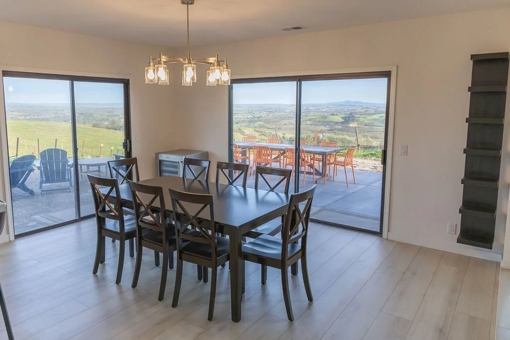 Dining room with black table and eight chairs, large sliding glass doors opening to a patio with outdoor dining table and chairs, scenic view of countryside with rolling hills and fields.
