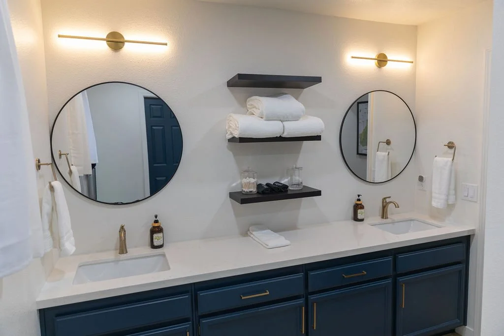 Bathroom with two round mirrors above a double sink vanity with navy blue cabinets and gold hardware, shelves with towels and toiletries, and wall-mounted lights.