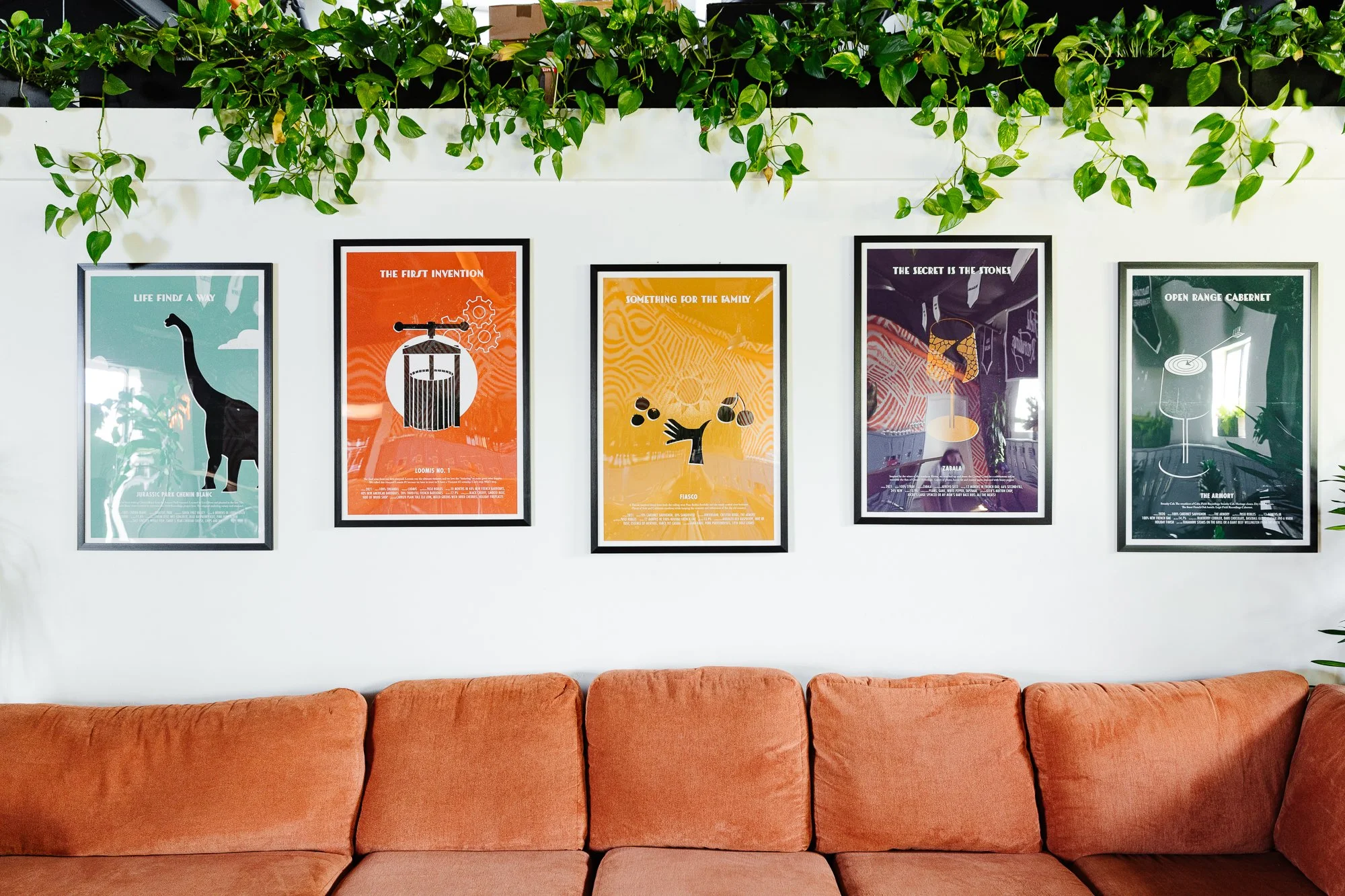Framed posters hanging on a white wall above an orange sofa, with green leafy plants hanging overhead.