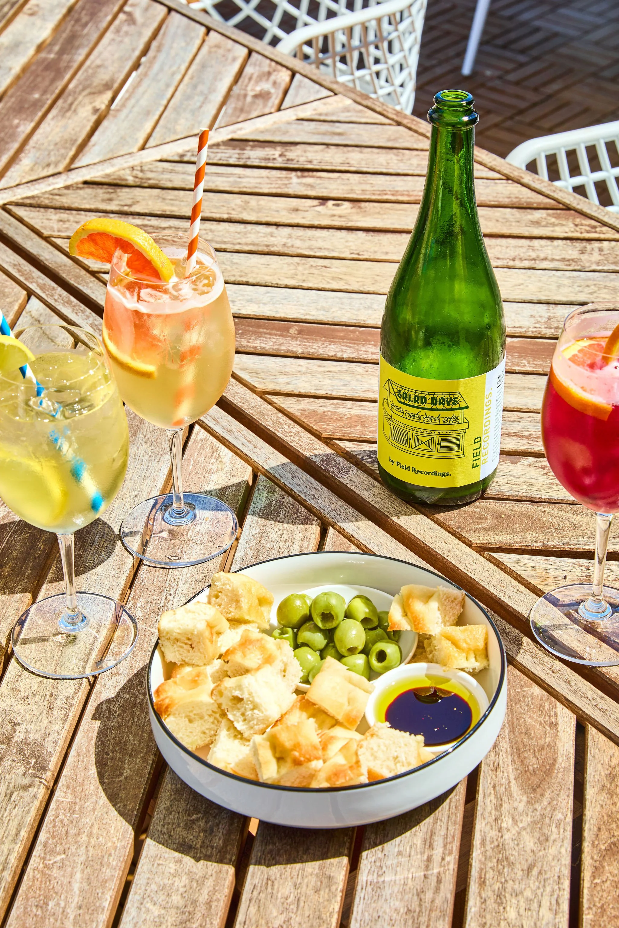 A wooden outdoor table with various drinks and snacks including a bottle of sparkling wine, colorful cocktails, a bowl of green olives, bread, and a small dish of balsamic and oil, with white chairs around.