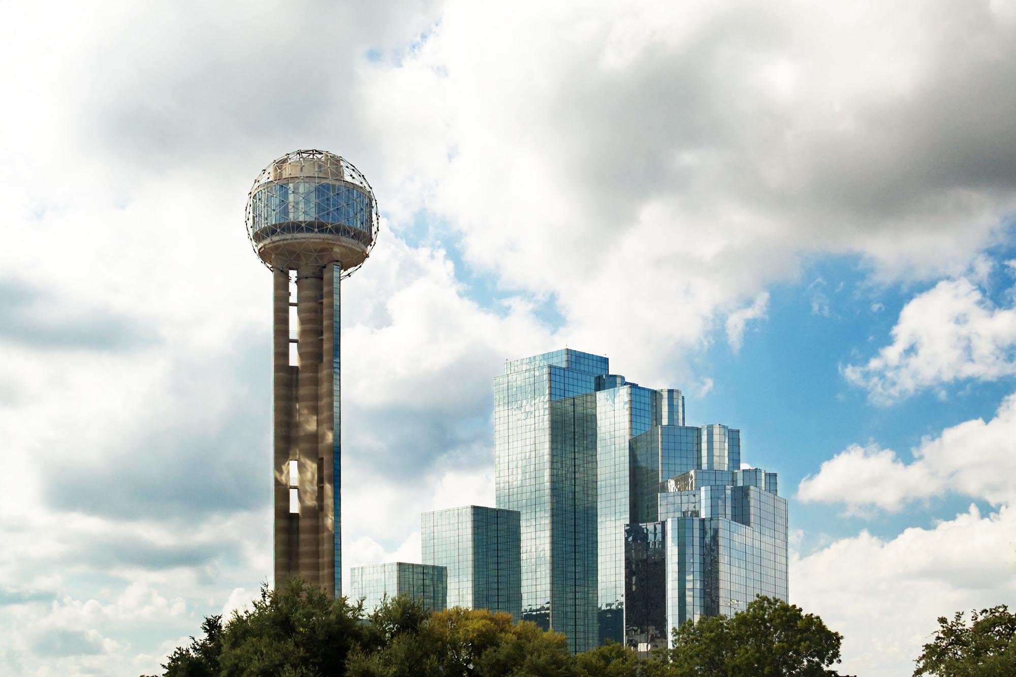 A city skyline featuring a tall observation tower with a spherical top made of glass and metal panels, along with modern glass office buildings reflecting the sky and clouds, with trees at the bottom of the image.