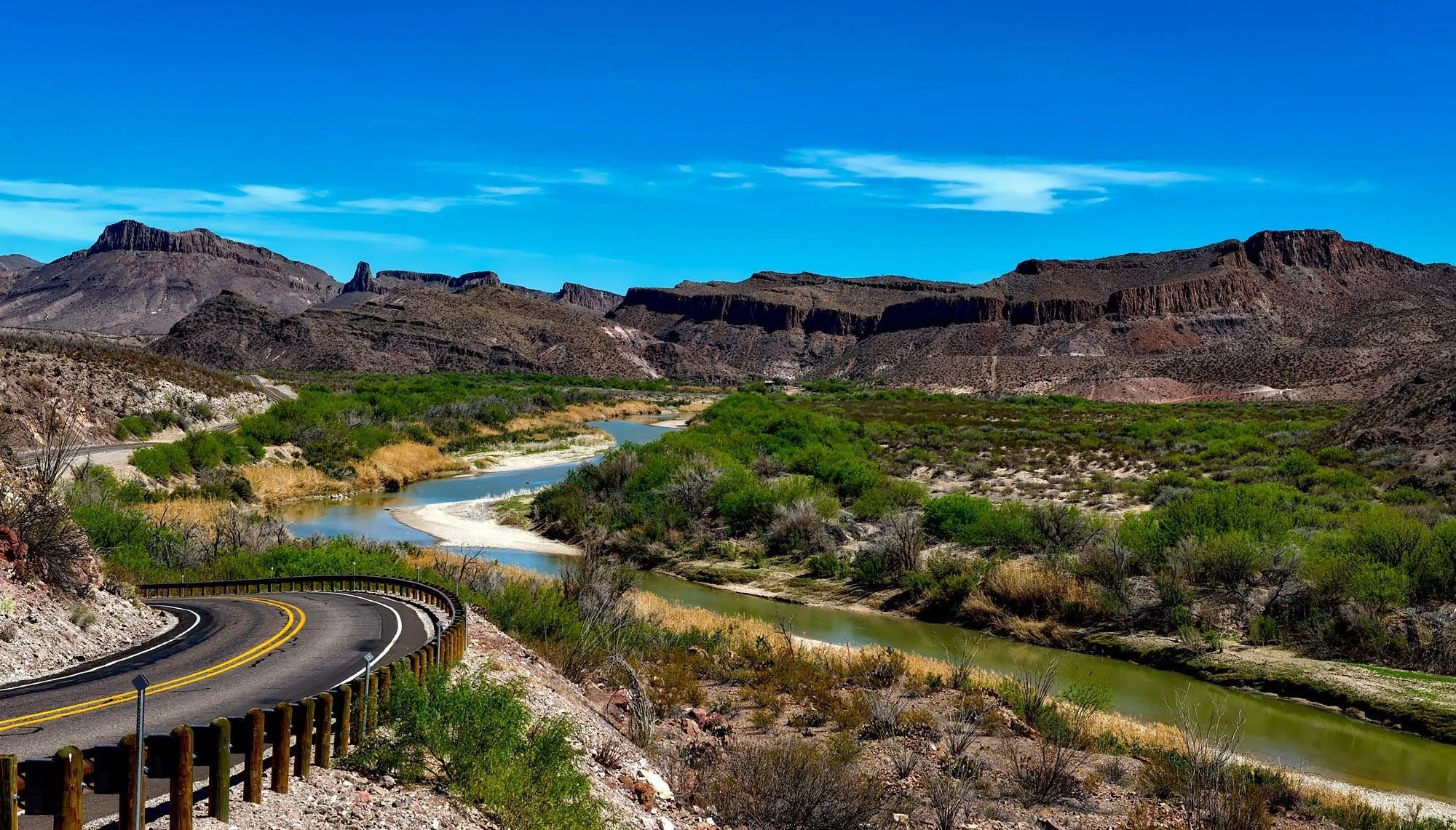 A winding two-lane road with a wooden guardrail runs along a green river in a desert landscape with rocky cliffs and mountains under a bright blue sky with a few clouds.