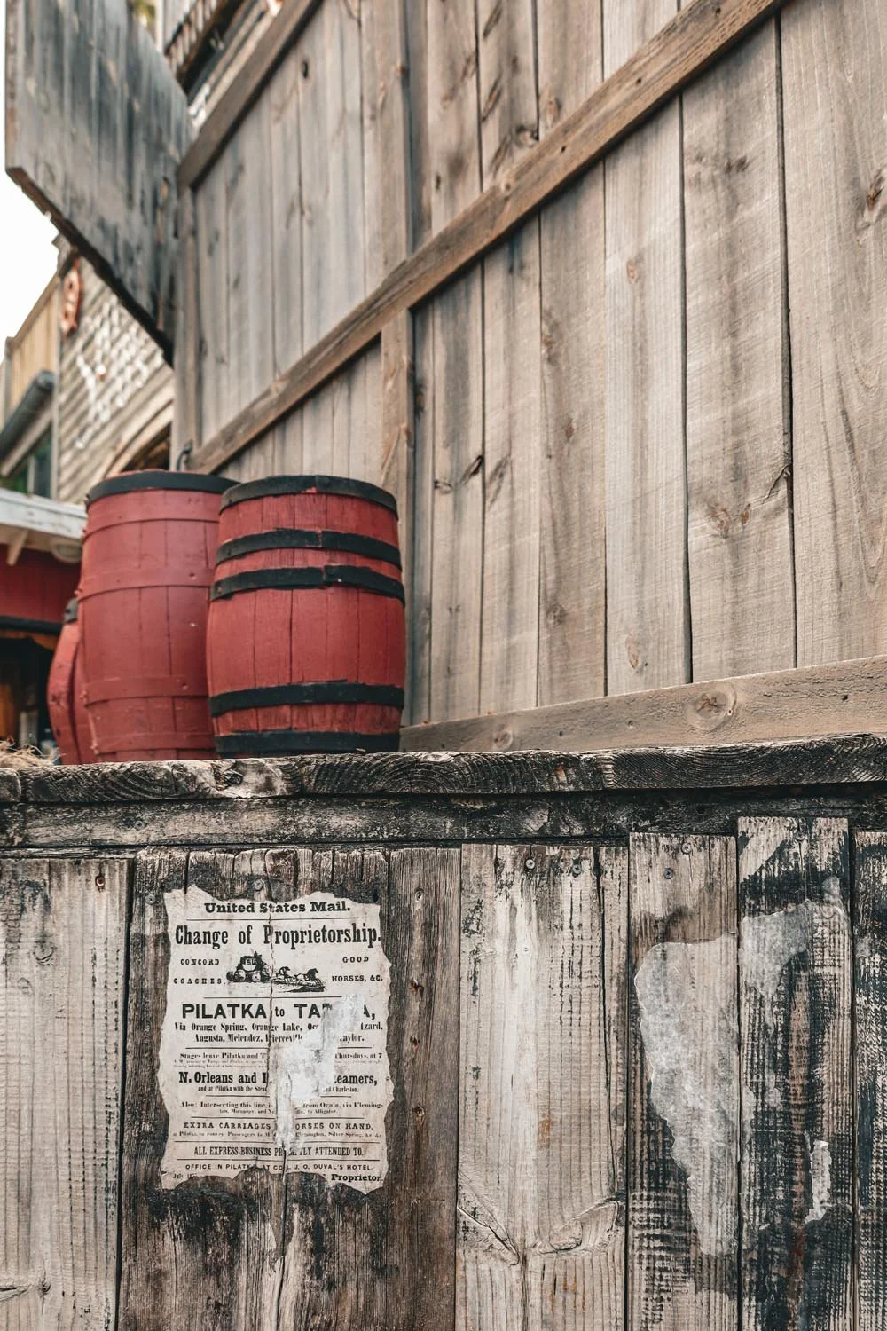 Two red wooden barrels with black metal bands on a weathered wooden platform in front of a tall, weathered wooden fence.