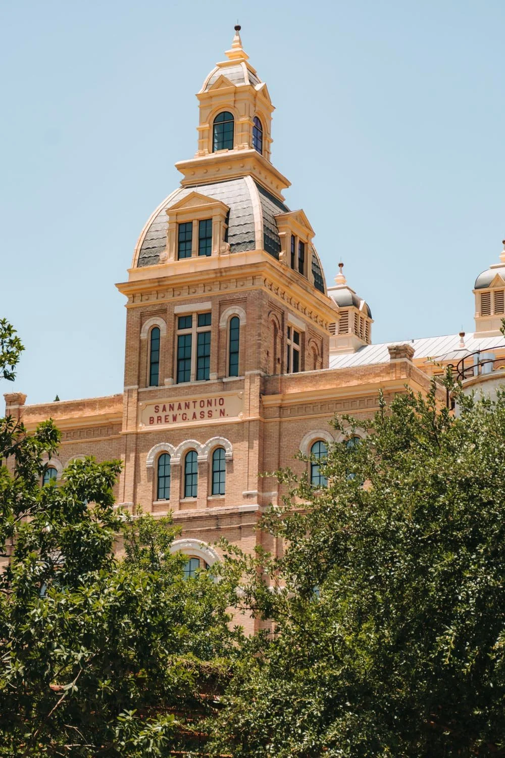 A historic building with a tall clock tower, called San Antonio Brew W. Gass N, surrounded by trees and set against a clear blue sky.