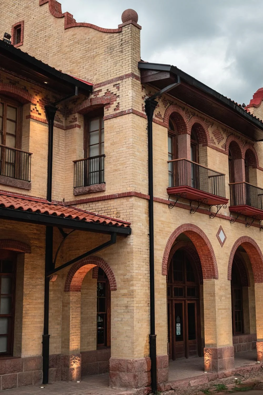 Close-up of a vintage brick building with arched windows and small balconies, black drainpipes, and a decorative brick pattern, under a cloudy sky.