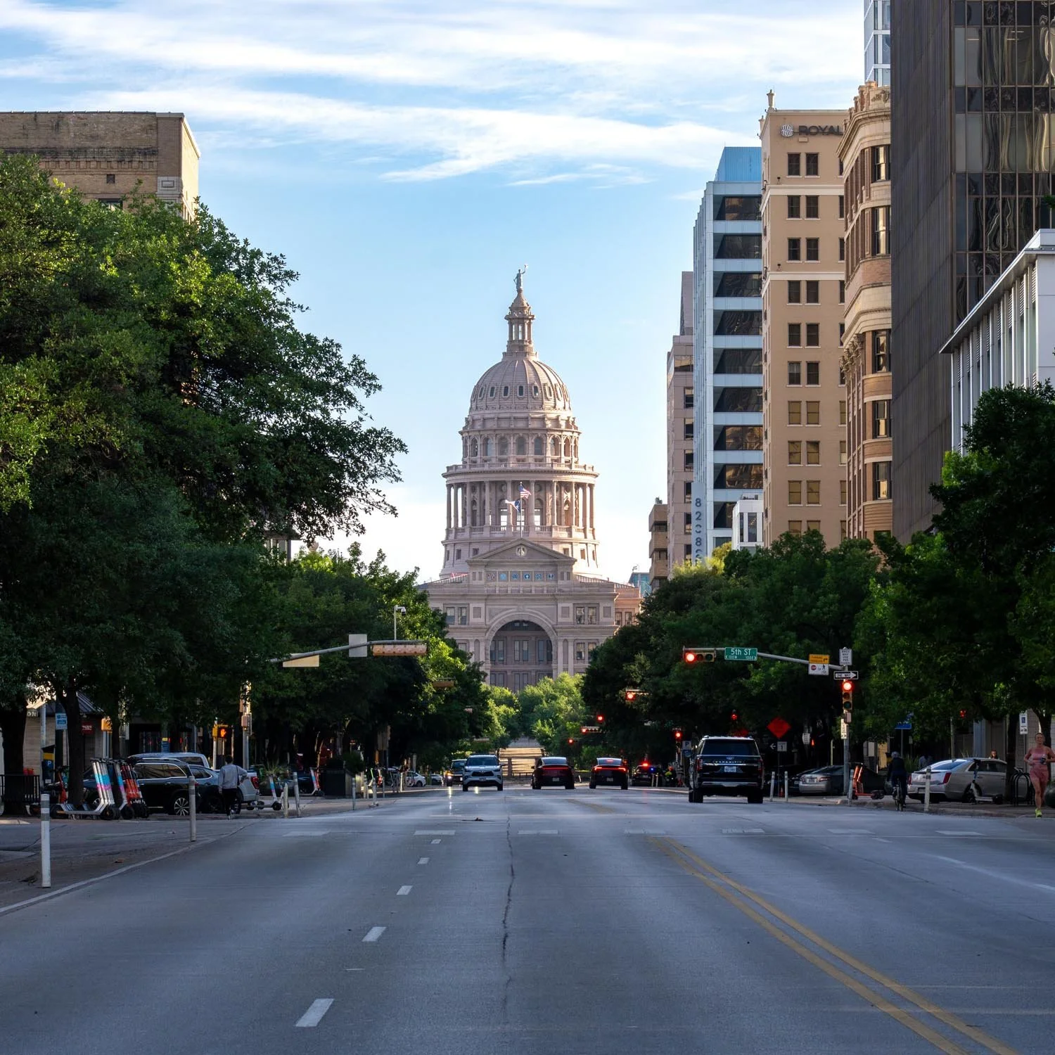 A city street view with trees lining the sidewalk, several cars parked and driving, and the Texas State Capitol building in the background under a partly cloudy sky.