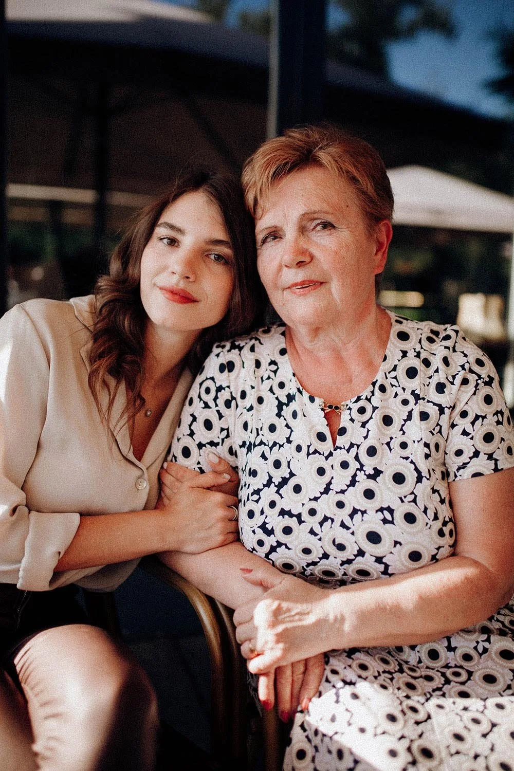 Two women, one elderly and one young, sitting close together outdoors, smiling and posing for the photo, with greenery and a canopy in the background.