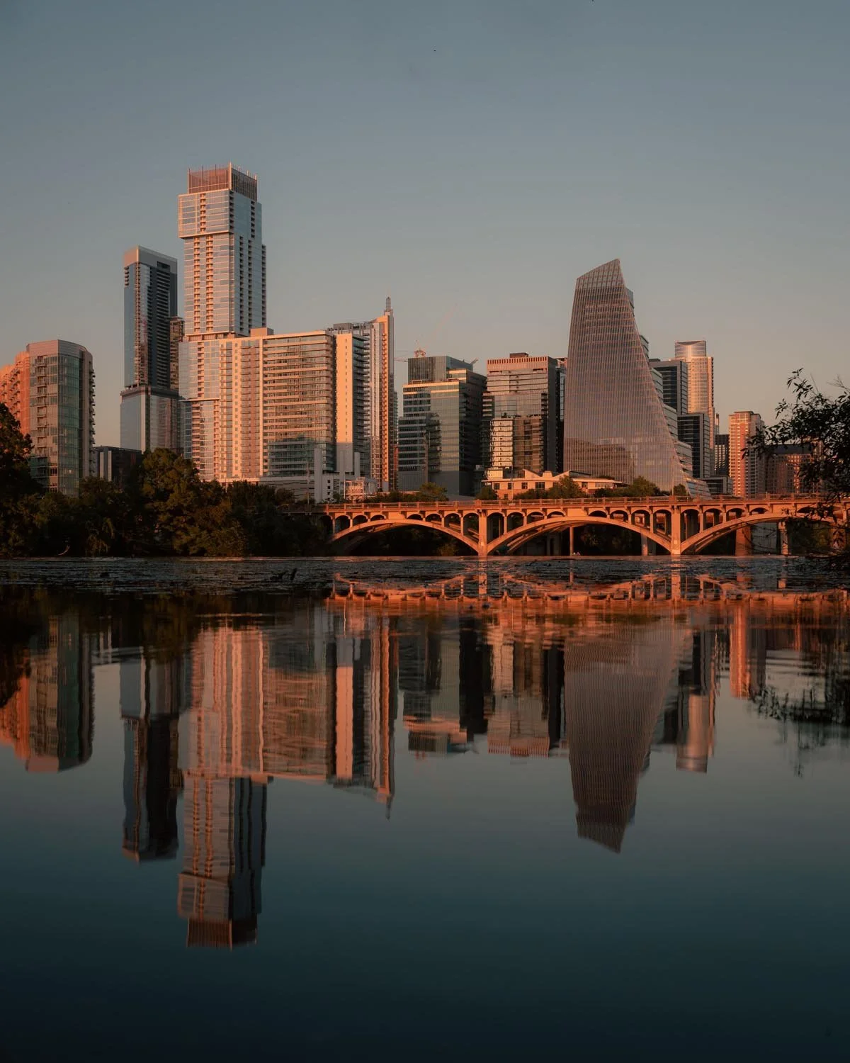 Sunset view of a city skyline with tall modern buildings reflecting on a river, featuring a bridge at the river's edge.