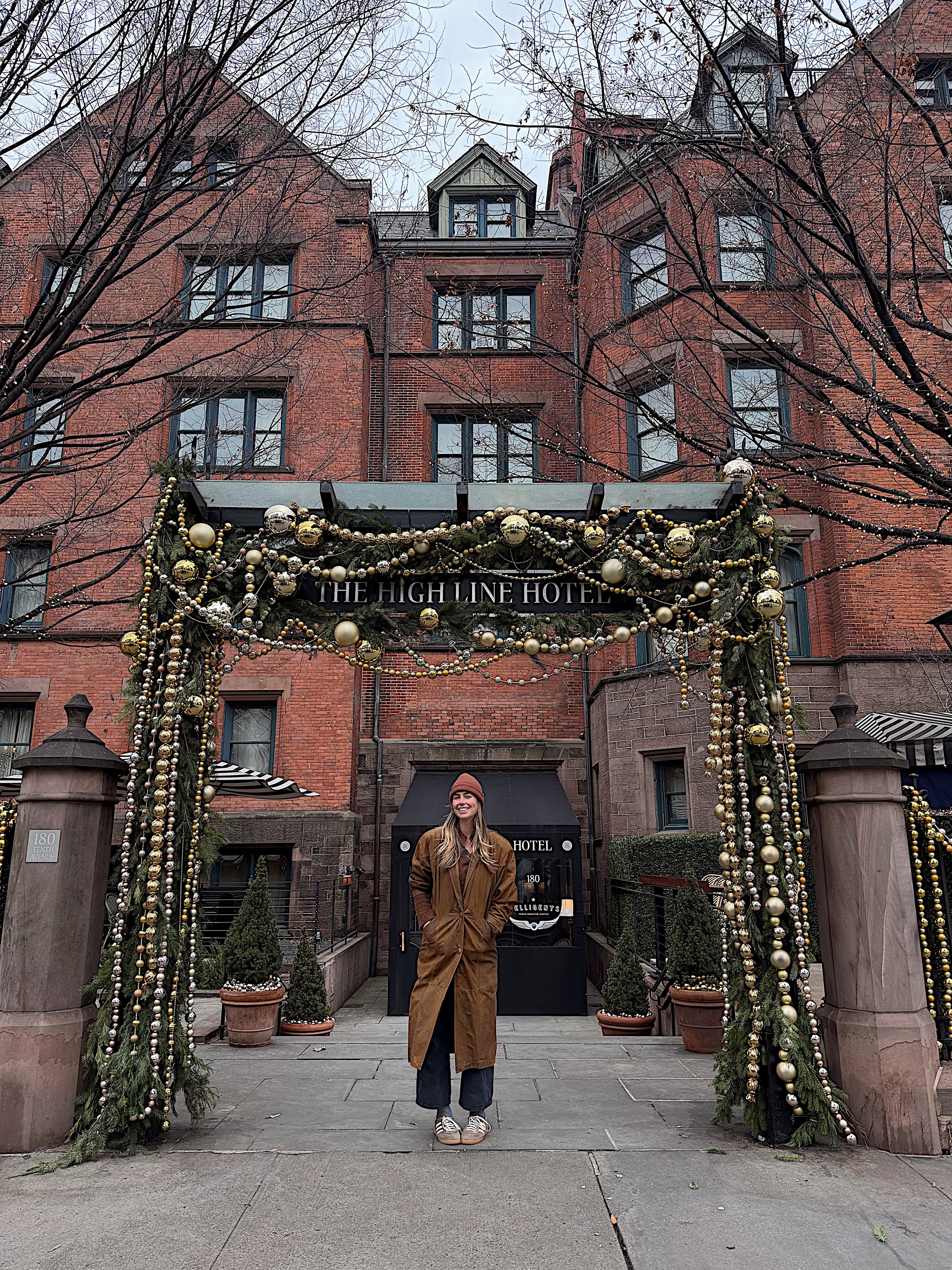Woman in brown coat and pink beanie standing at the entrance of The High Line Hotel, decorated with Christmas ornaments and greenery, in front of a brick building.