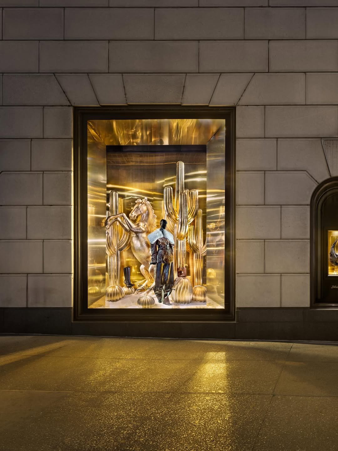 Luxury retail window display with a golden horse, cacti, gold domes, and mannequins dressed in fashionable clothing, illuminated with warm lighting.
