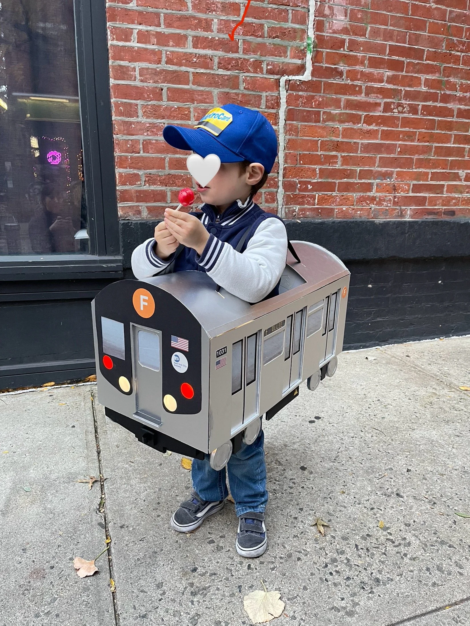 A young boy wearing a blue baseball cap and a jacket with striped sleeves is standing outdoors on a concrete sidewalk wearing a custom subway train Halloween costume