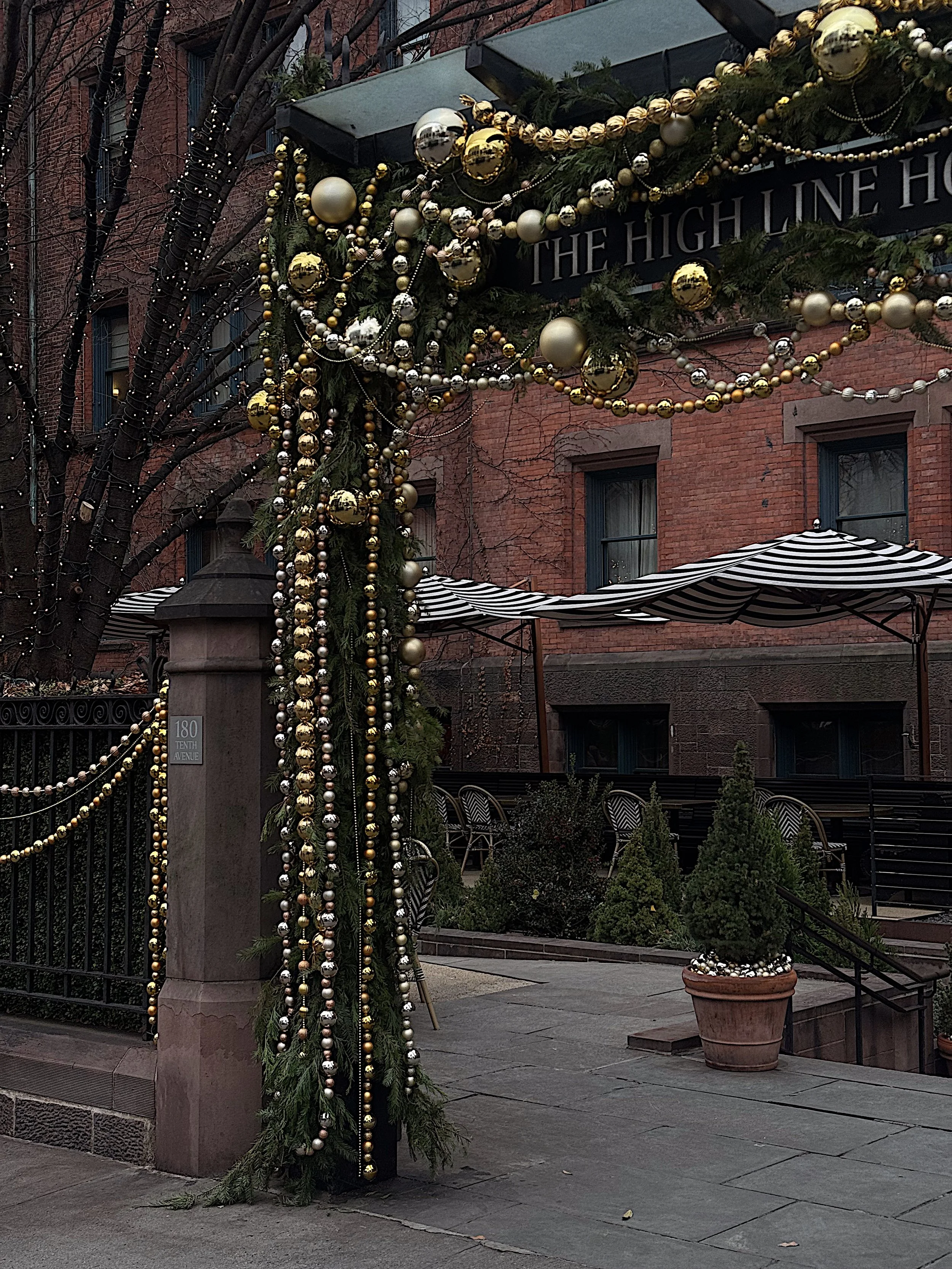Christmas decorations on a street corner outside a building, including garlands with gold and silver ornaments and bead strands, with potted plants and striped awnings in the background.
