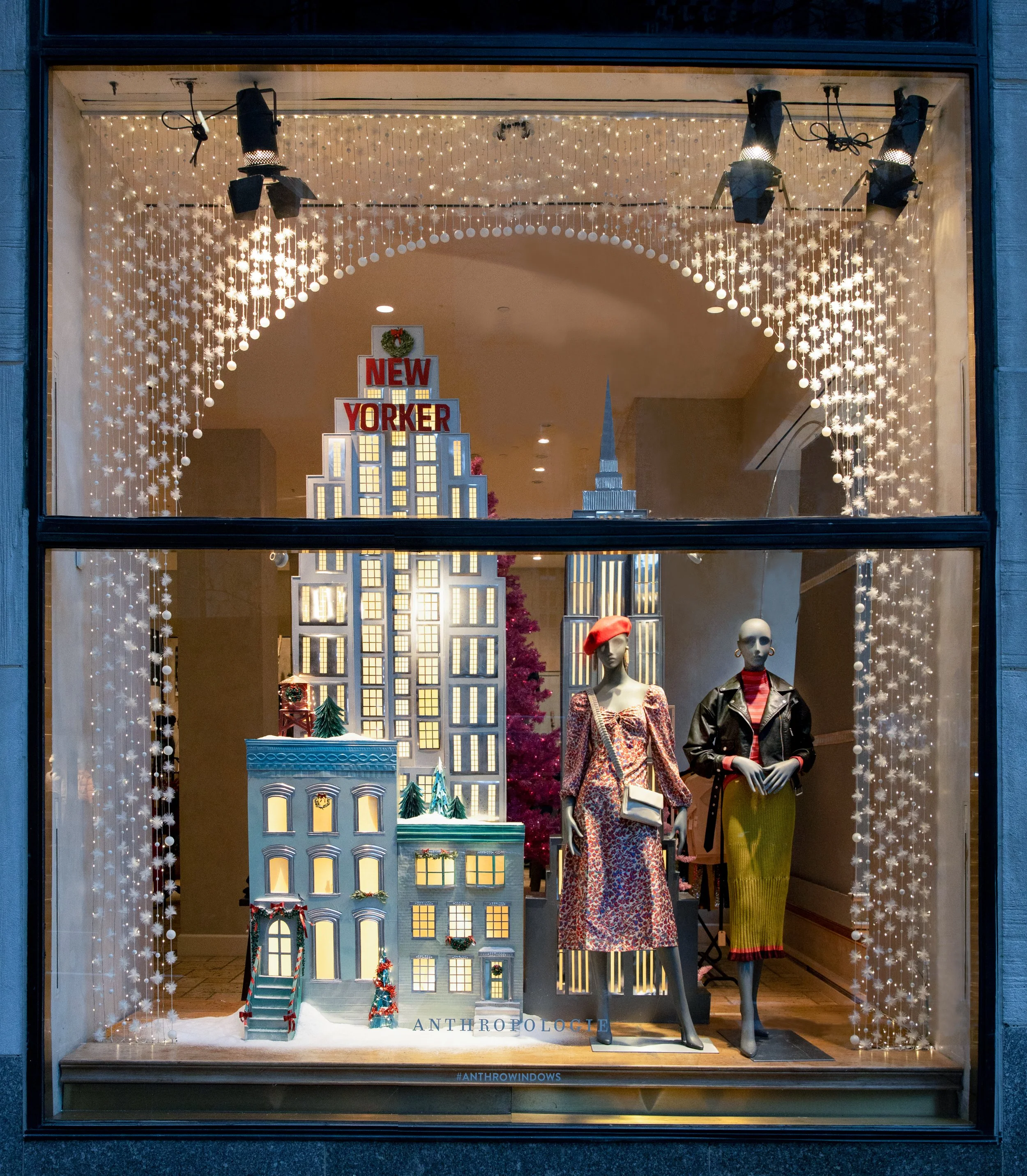 Holiday window display with Christmas decorations, including a miniature cityscape with New Yorker sign on a tall building, two mannequins dressed in fashionable winter clothing, and festive string lights framing the scene.