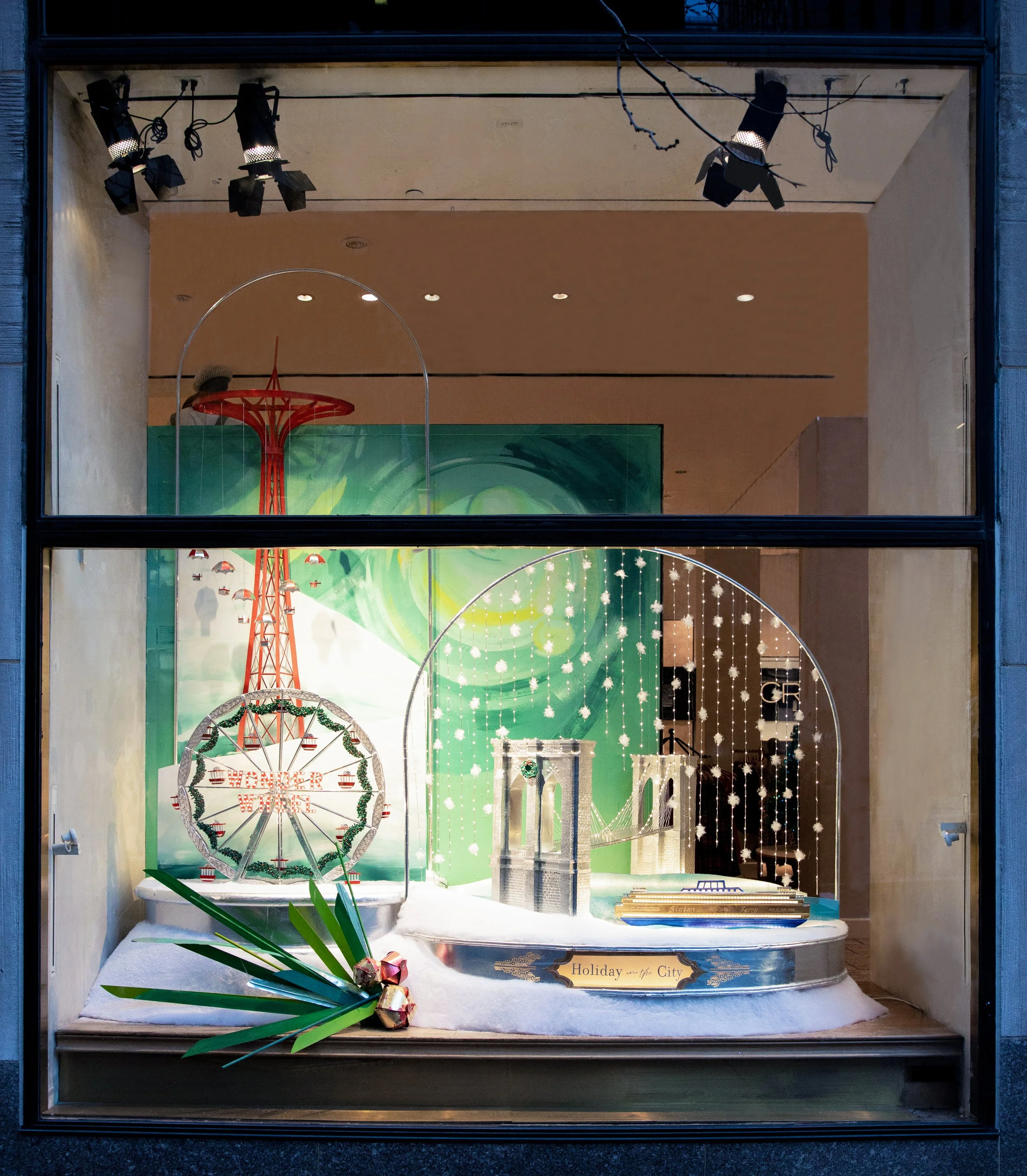 Holiday-themed window display with miniature Ferris wheel, false snow, a model bridge, and a green background with lights and decorations.