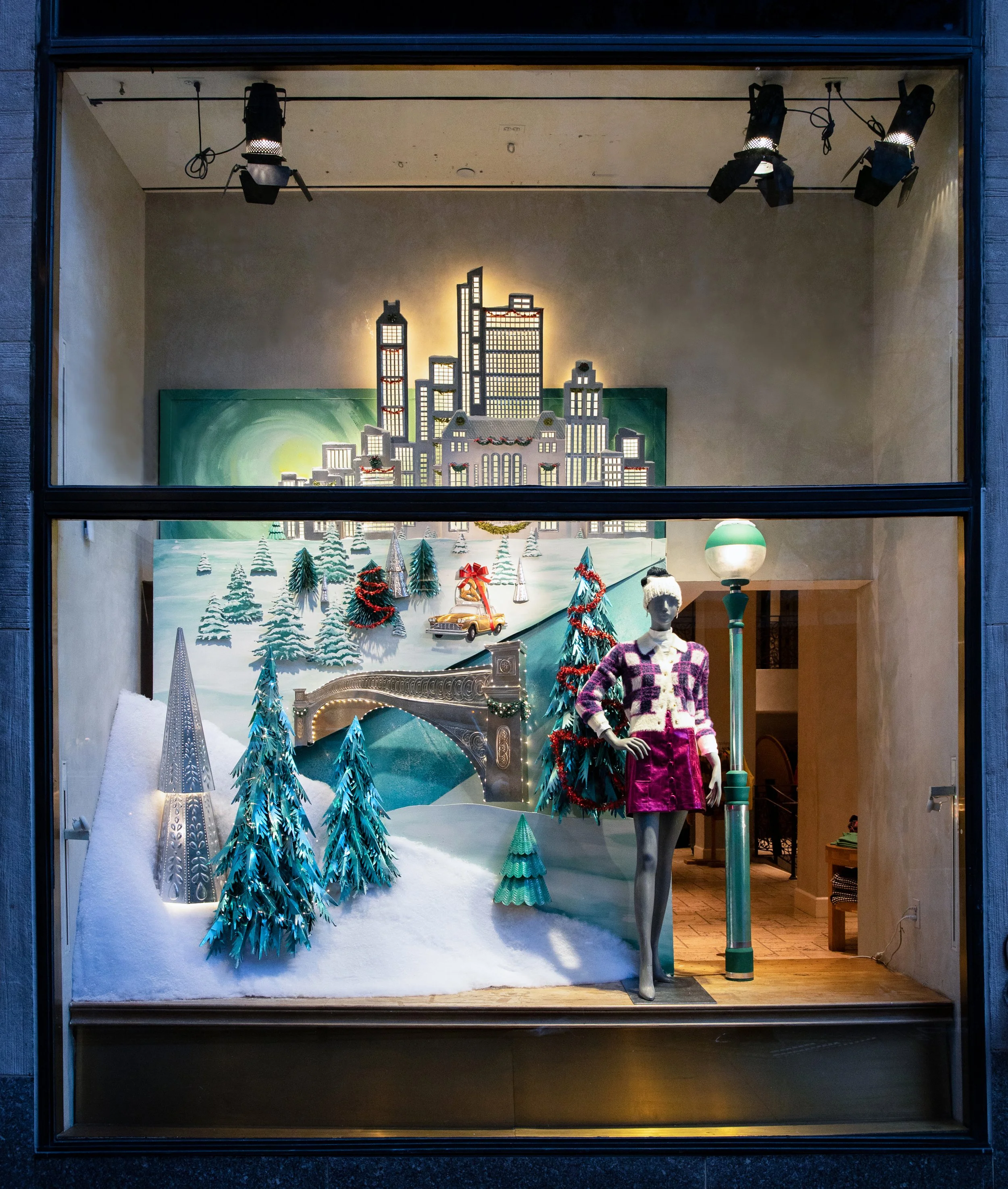 Holiday window display featuring a miniature winter scene with snow-covered trees, a small bridge, and vintage cars, set against a backdrop of a city skyline, with mannequin dressed in a purple and white sweater and a red skirt, and festive decorations including Christmas wreaths and lights.