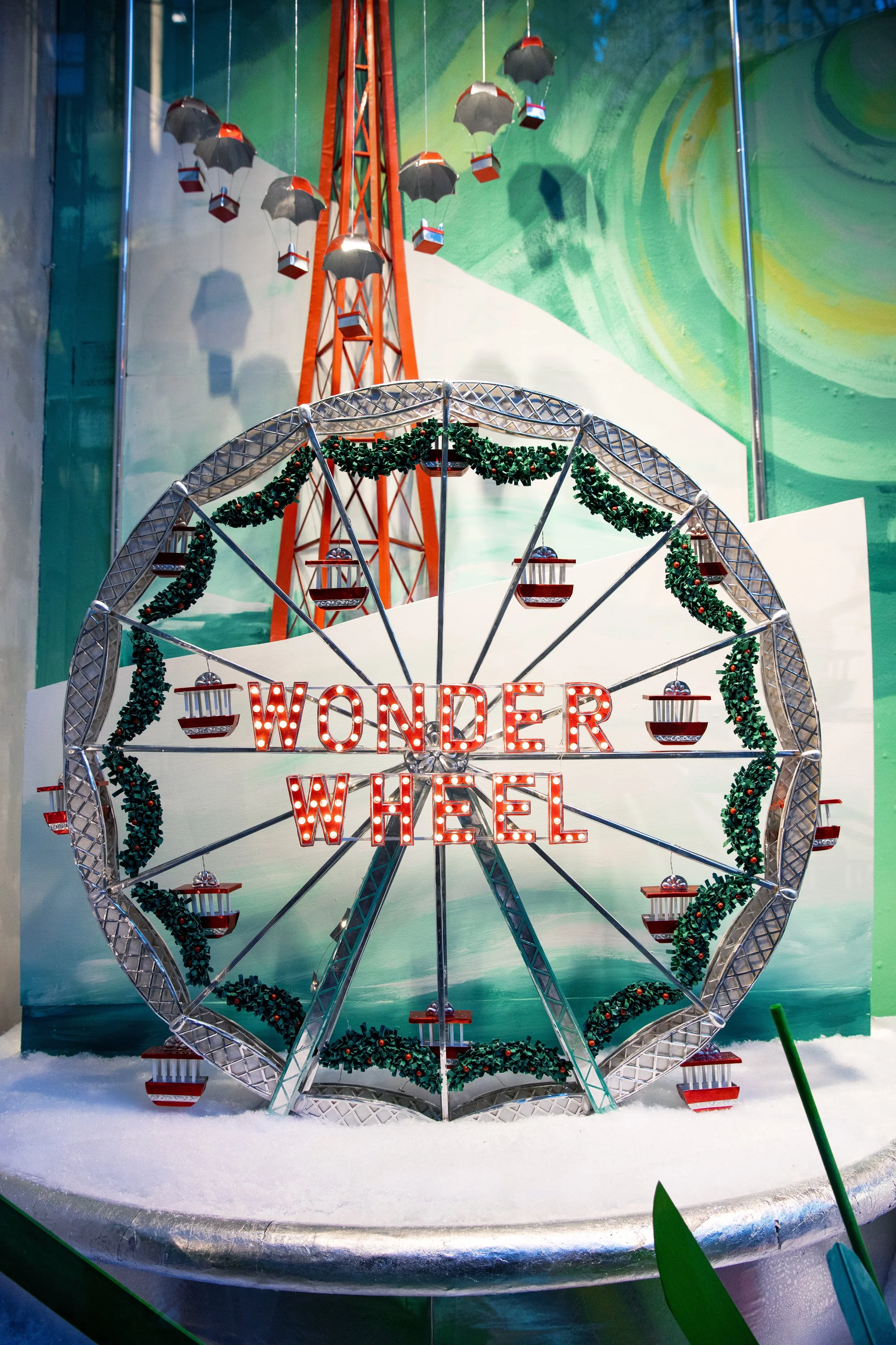 A festive amusement park decoration called Wonder Wheel with a large illuminated sign, decorated with green garlands, surrounded by snow, with a colorful backdrop and a tower in the background.