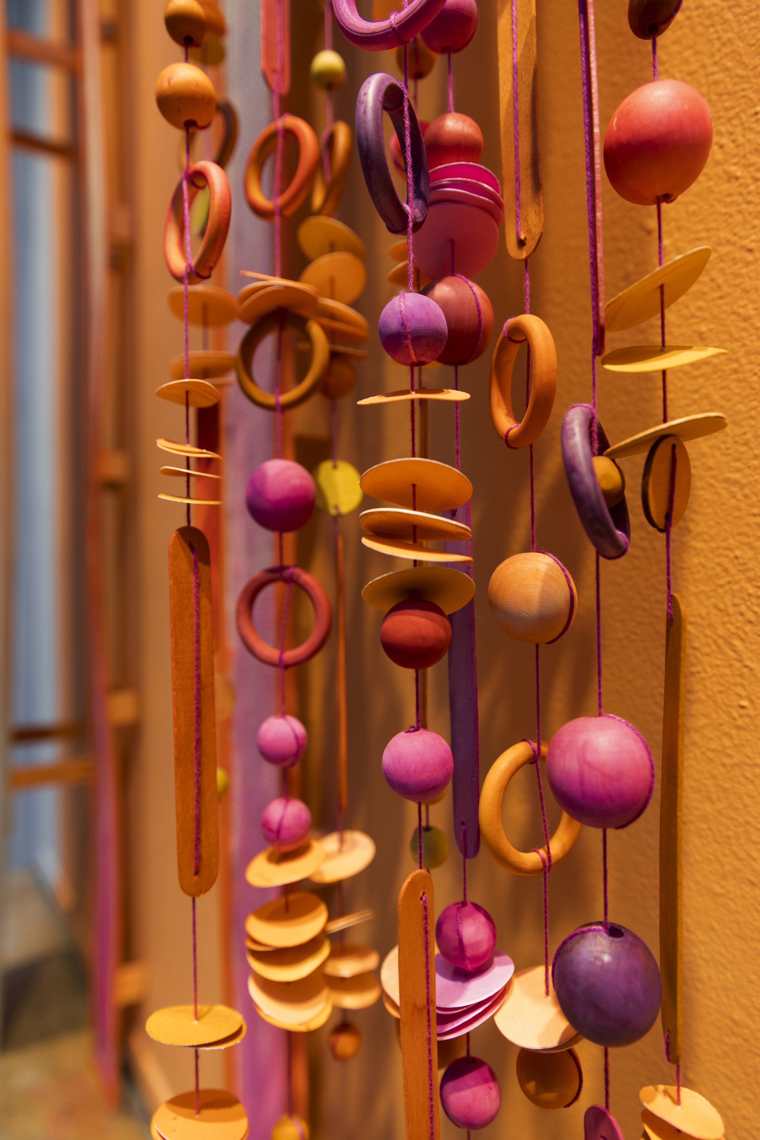 Colorful wooden hanging mobile with circular and spherical shapes in shades of pink, orange, purple, and yellow, hanging against a warm-colored wall.