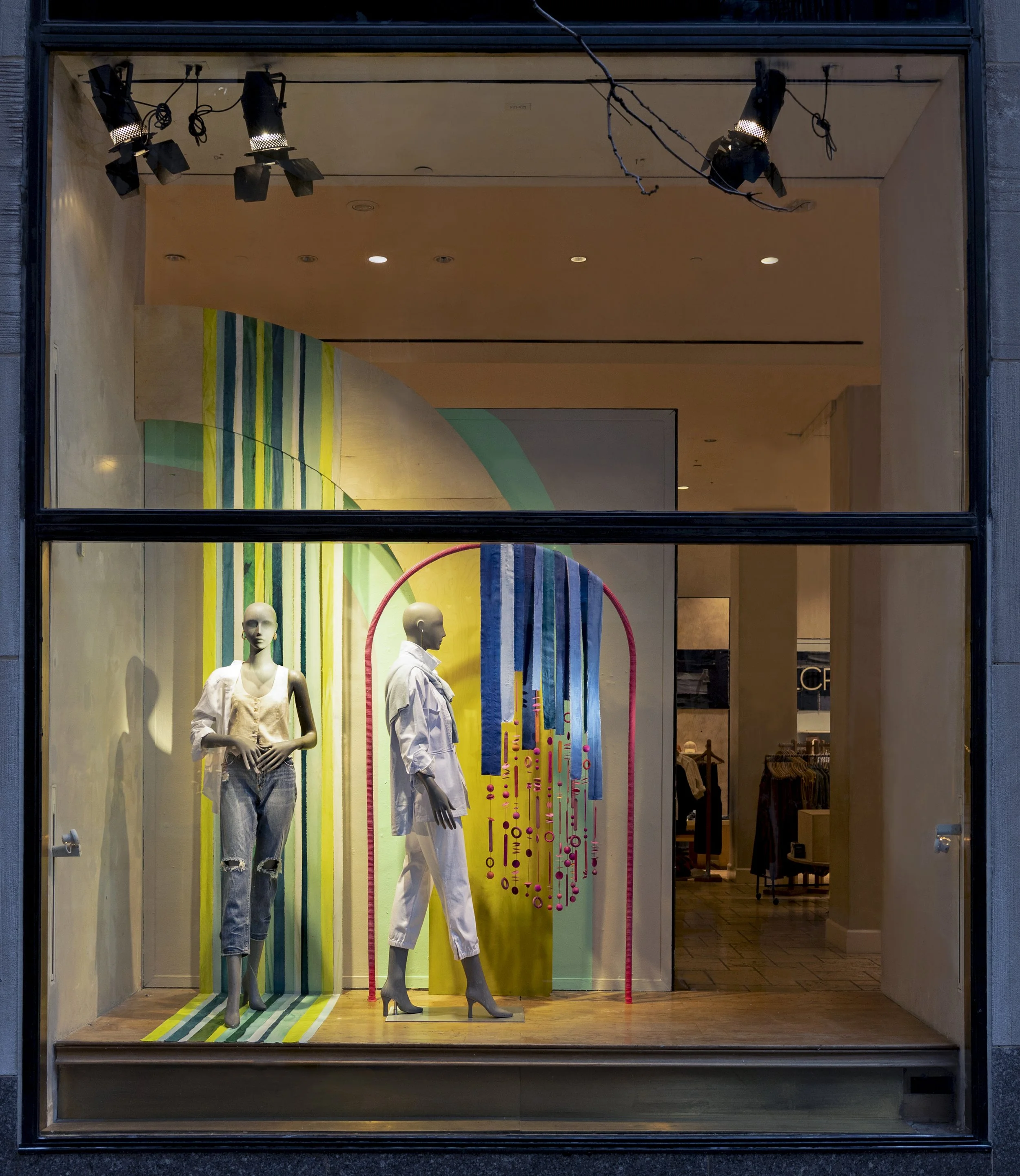 Store window display with mannequins dressed in casual outfits, colorful striped backdrop, abstract art with hanging blue and pink elements, and store interior in the background.