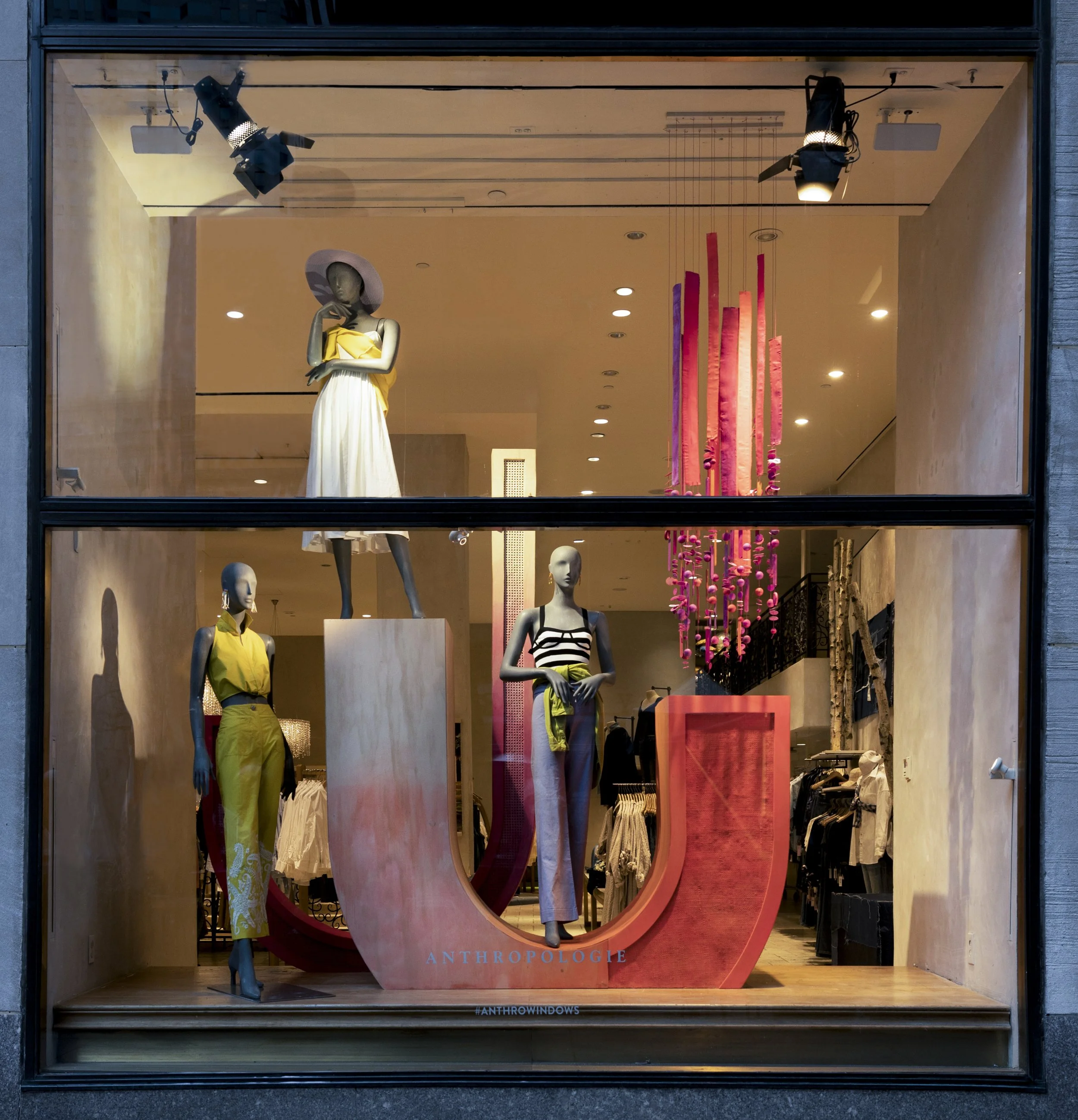 Display window featuring mannequins dressed in fashionable clothing, with a large coral-colored 'U' sign reading 'Anthropologie' and a pink chandelier hanging in the background.