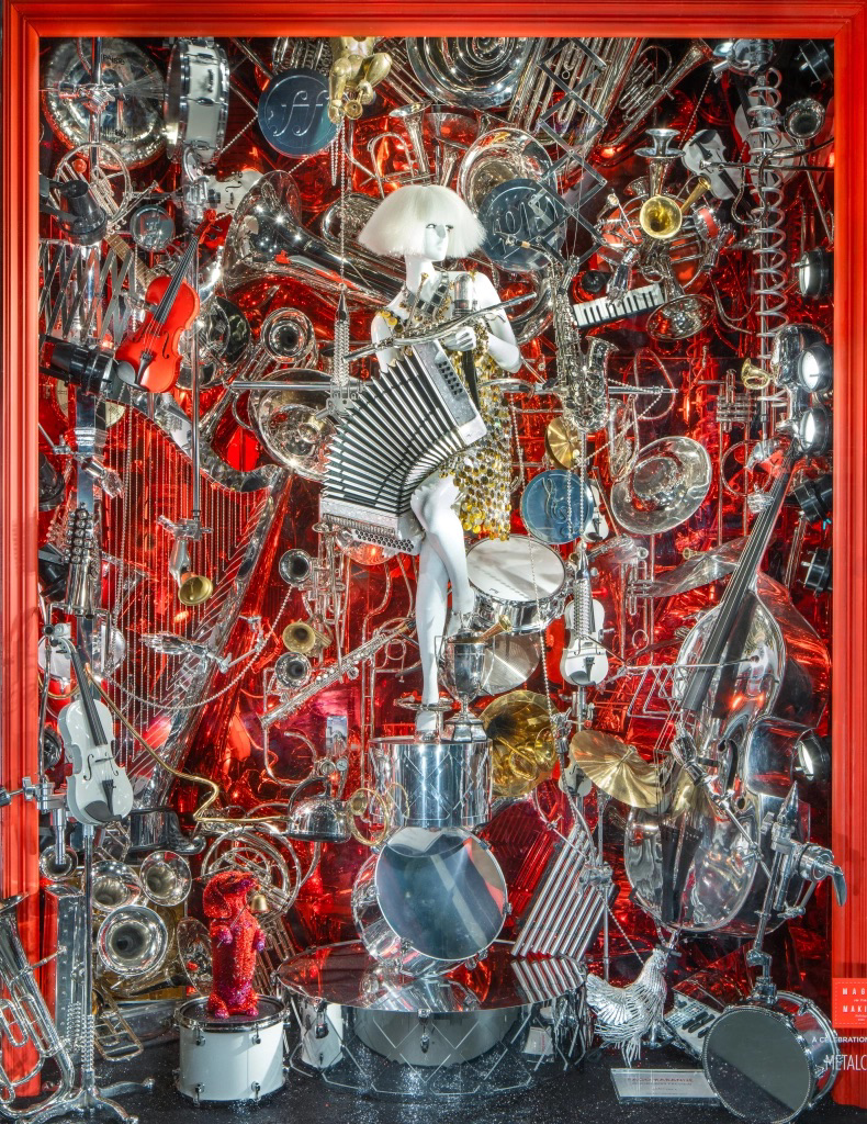 An elaborate display of musical instruments and decorations, including violins, drums, keyboards, cymbals, and metallic sculptures, with a central figure of a person with a large white wig playing a keyboard, all set against a shiny red background.