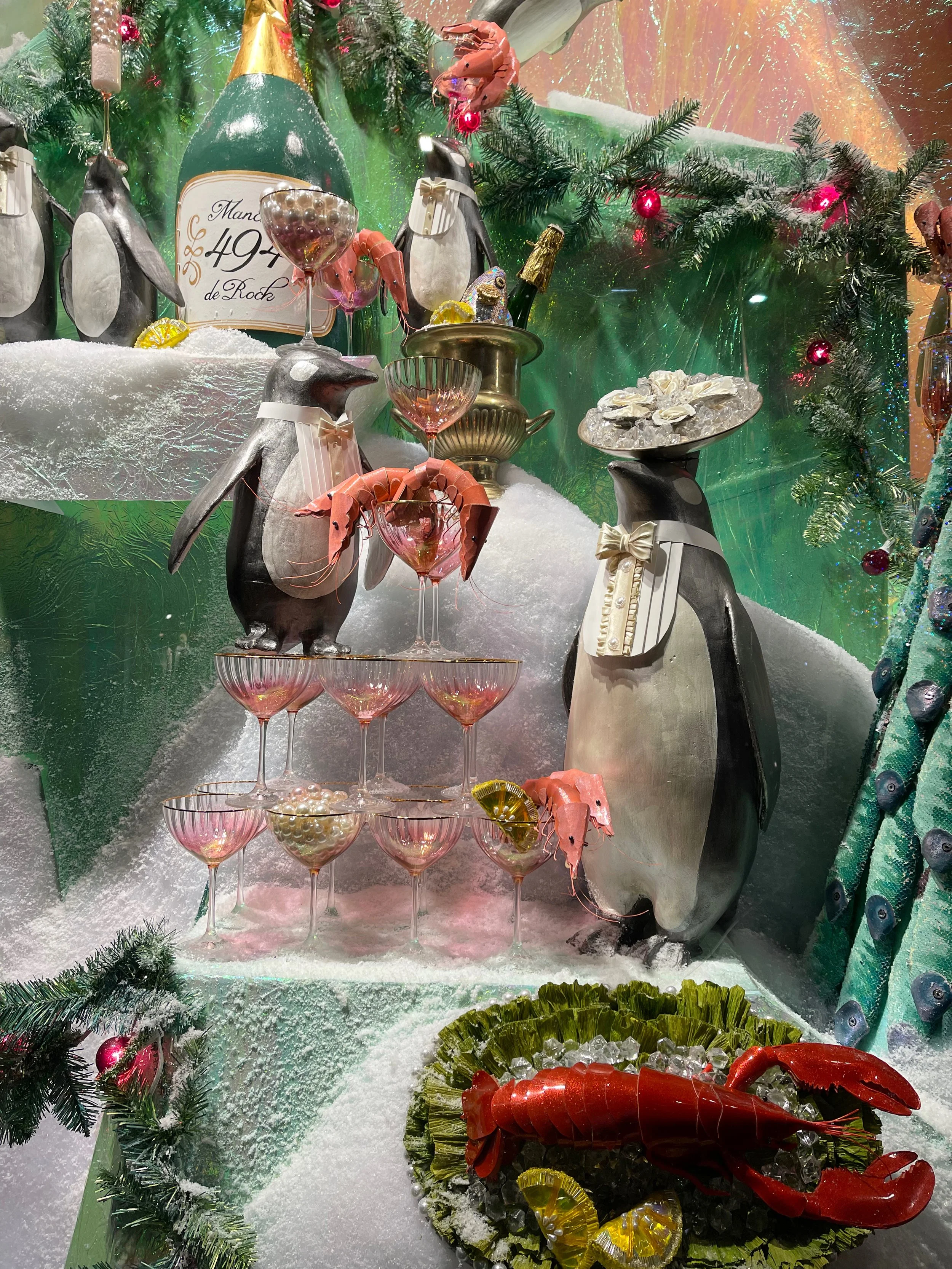 Festive display with penguins, champagne bottle, glasses, and seafood on an icy, snow-covered surface decorated with Christmas greenery and ornaments.