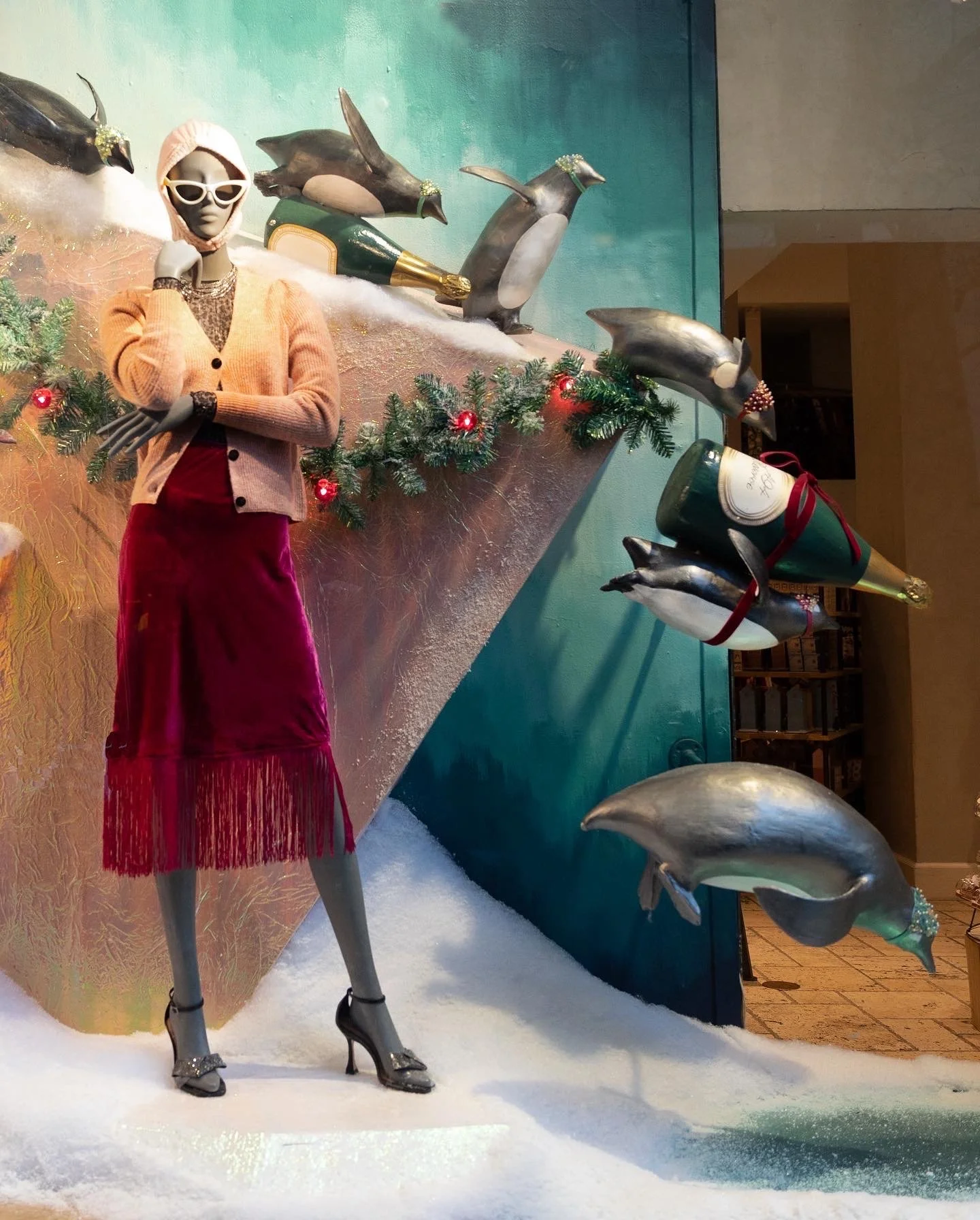 Holiday mannequin display with a female figure wearing a beige cardigan, red skirt, and high heels, surrounded by penguin decorations and festive greenery.