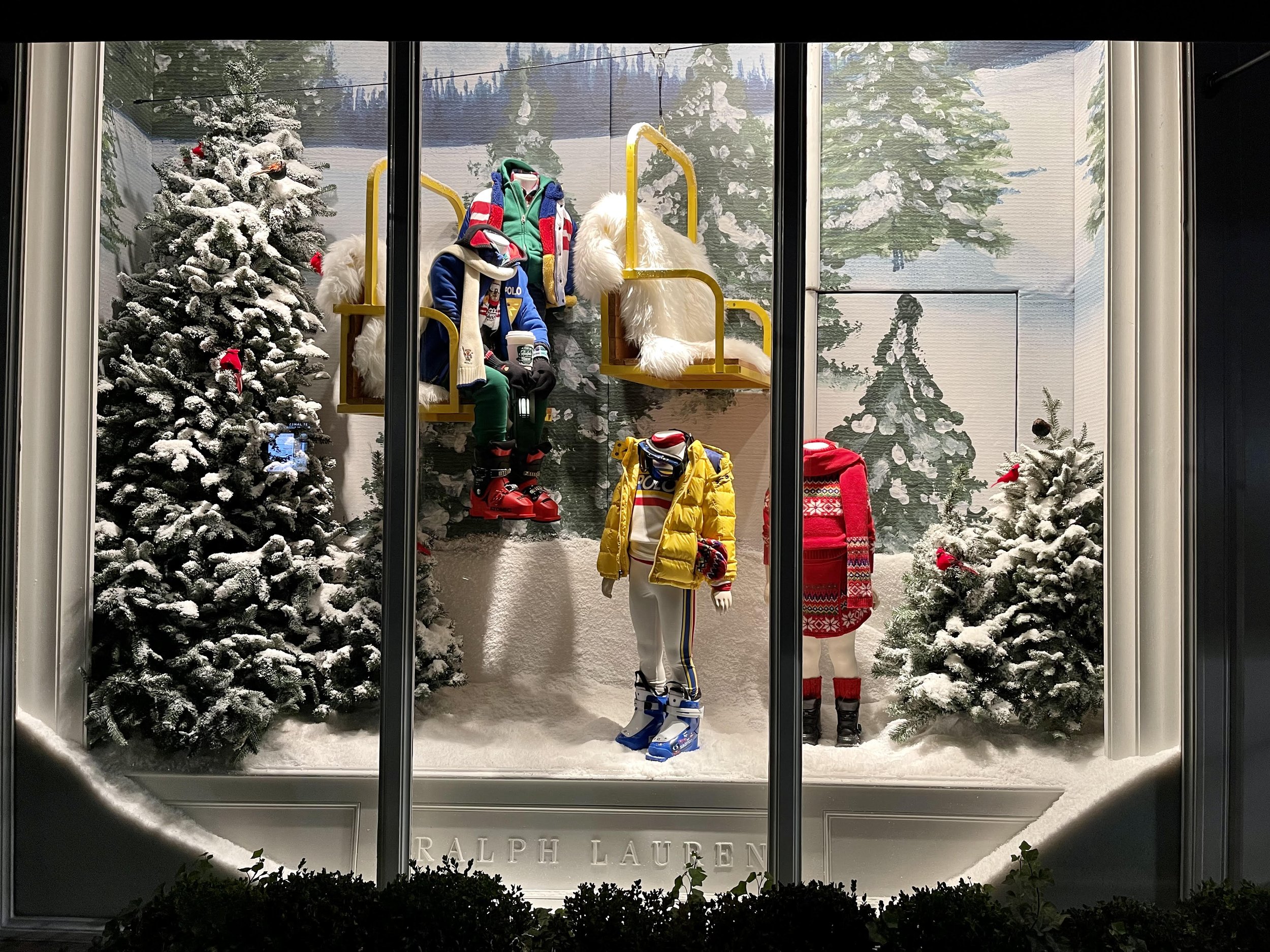 Display window with winter clothing and accessories including mannequins dressed in winter outfits, snow-covered trees, and a snowy landscape background, featuring the Ralph Lauren brand name.